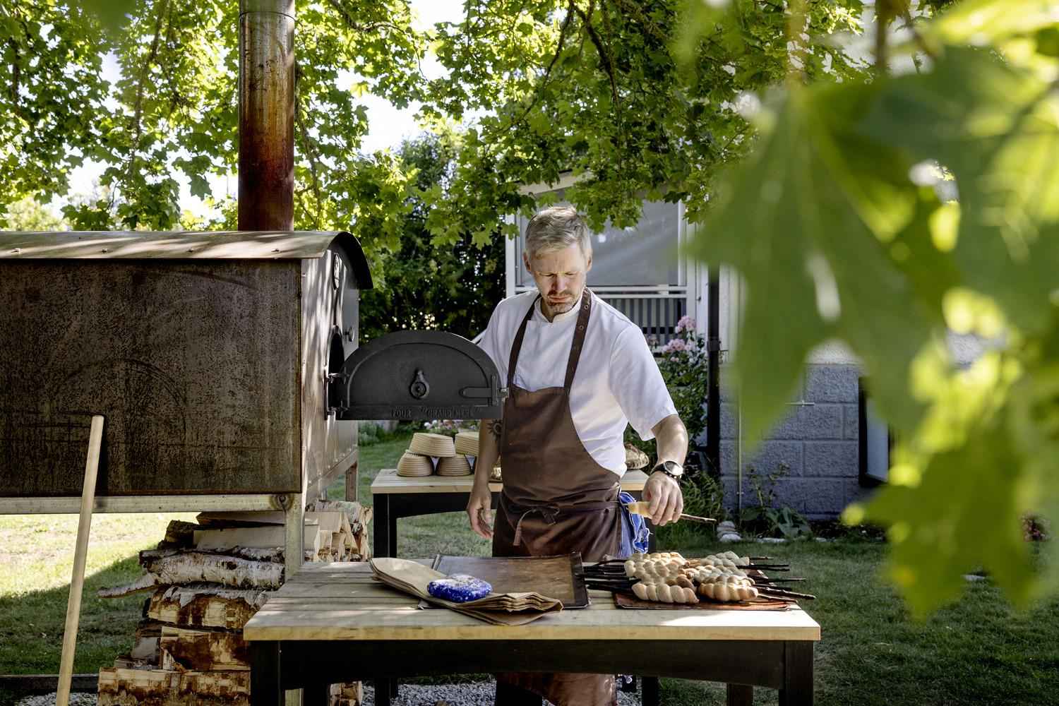 A chef is standing by a wood fired oven in a garden.