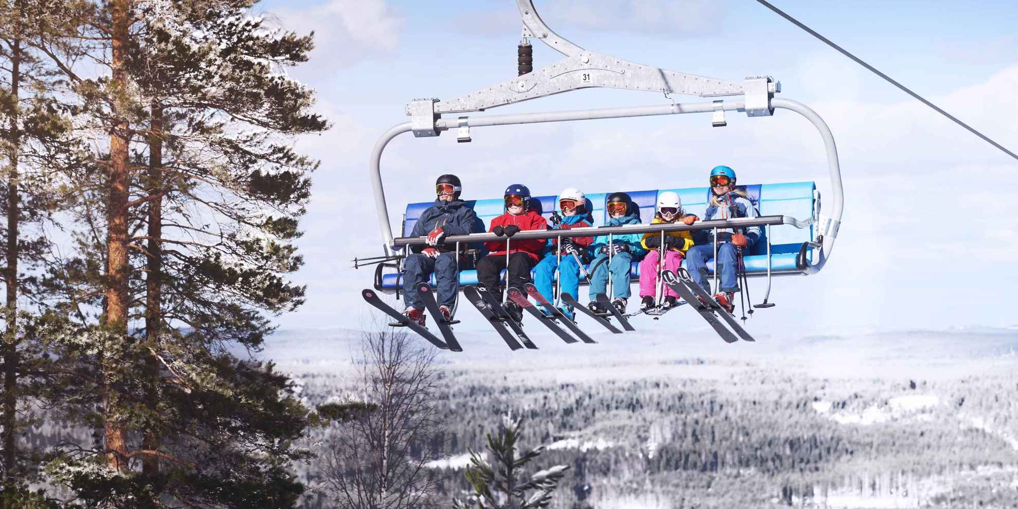 A family of six people sits in a ski lift with ski gear going up a hill during winter.