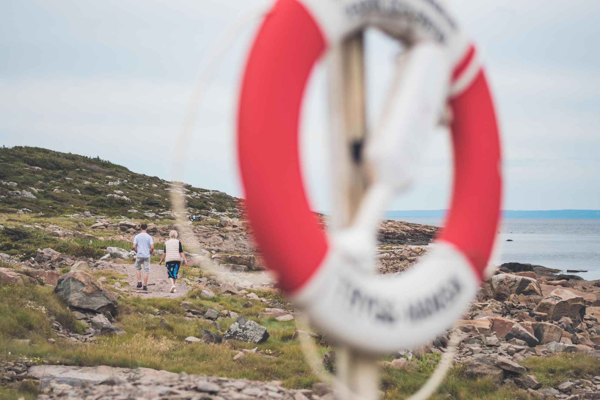 A couple walks on cliffs at a coastline trail. A blurry lifebuoy in the front.