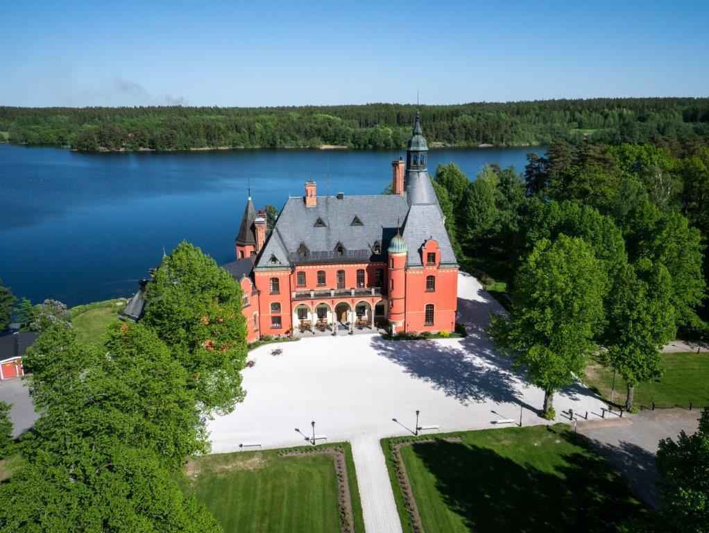 A castle with a red facade is surrounded by greenery and a lake.