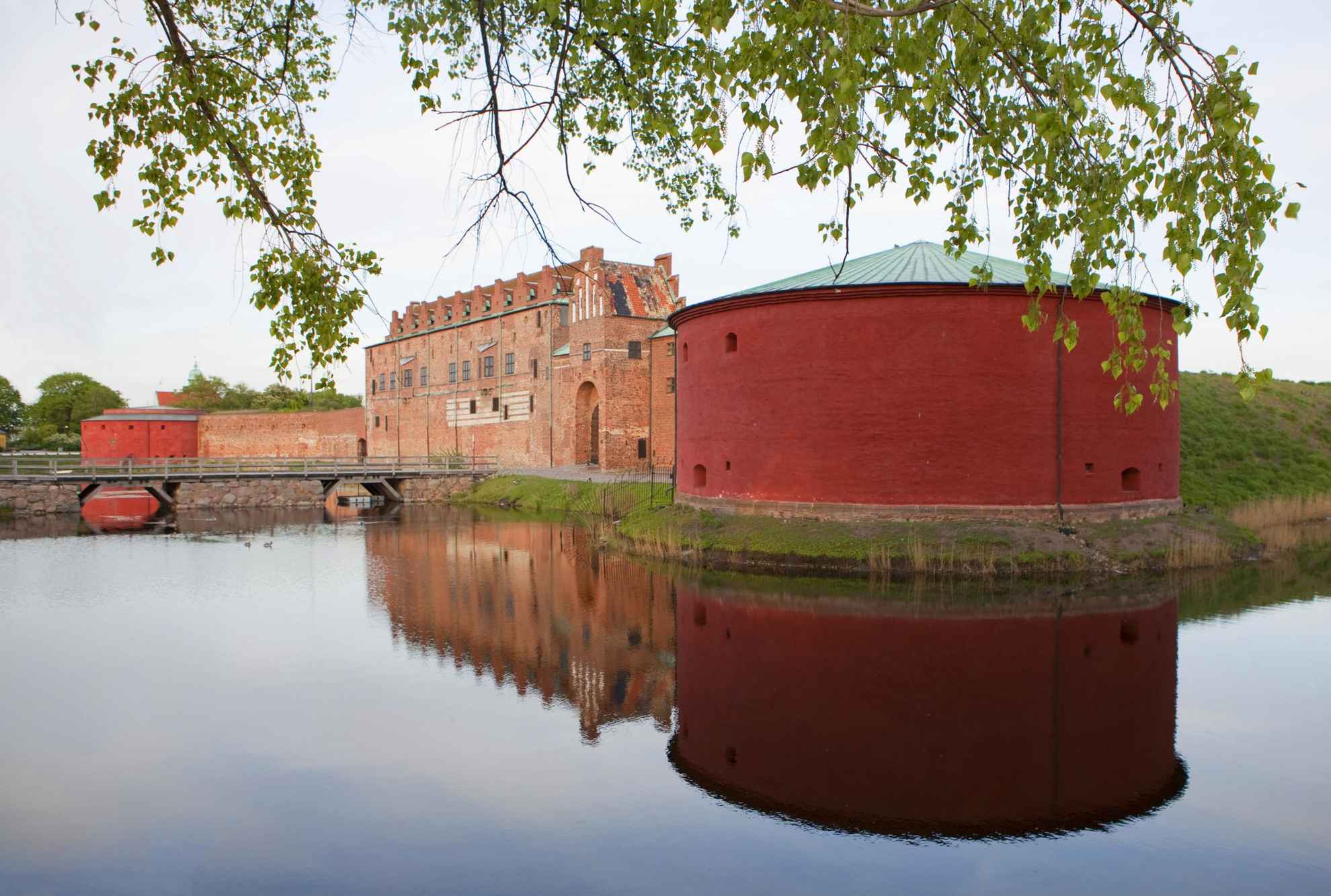 Malmöhus Castle is a large round tower next to a brick building, surrounded by water. Another round tower on the opposite side in the background. Green leaves in the foreground.