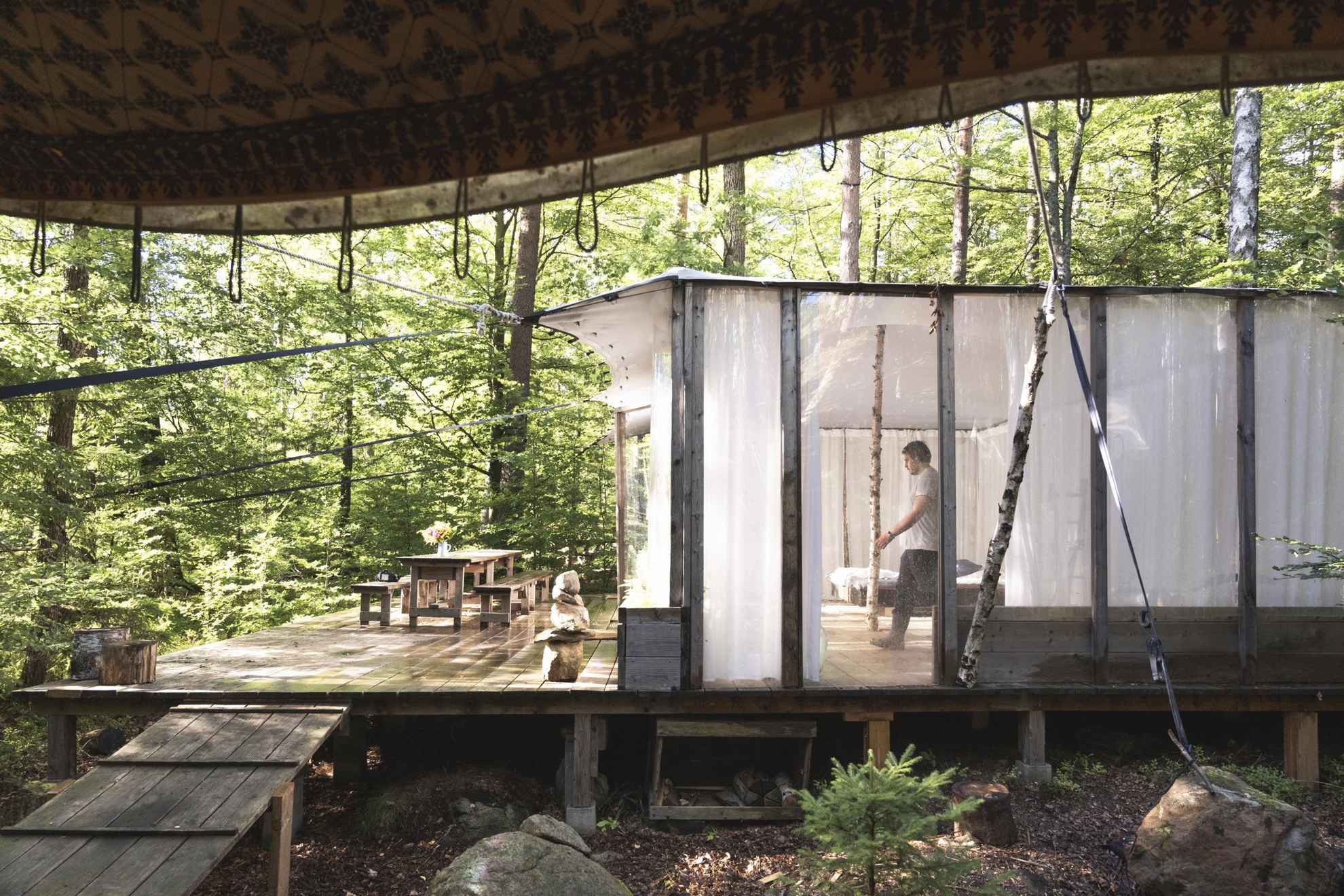 Exterior of a light and transparent tree-house style cottage in the woods, with green foliage all around and a man walking through the room.