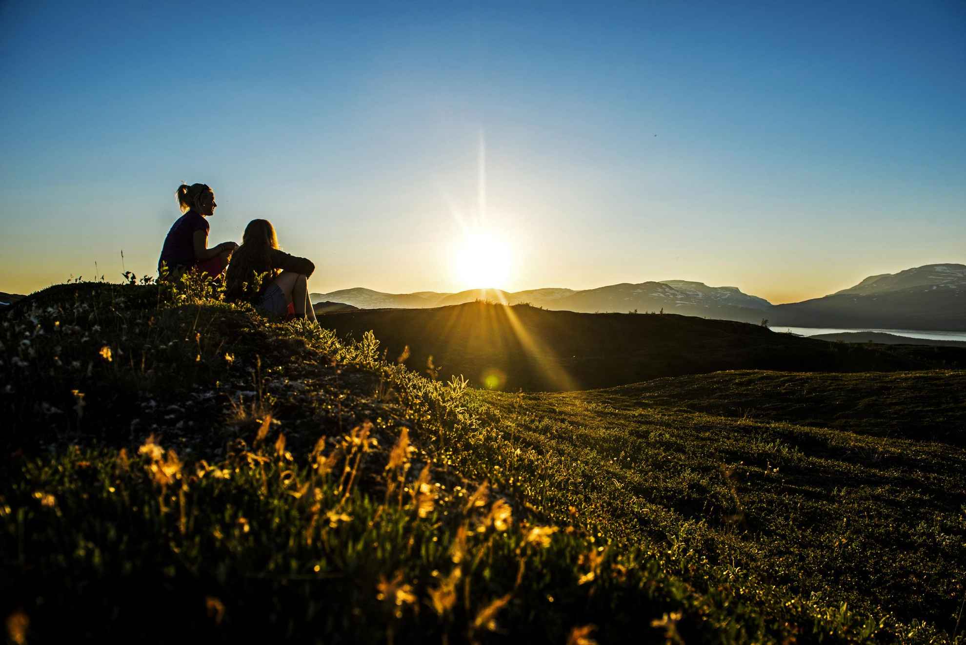 Two women are sitting on a hill in the mountains and enjoying the midnight sun.