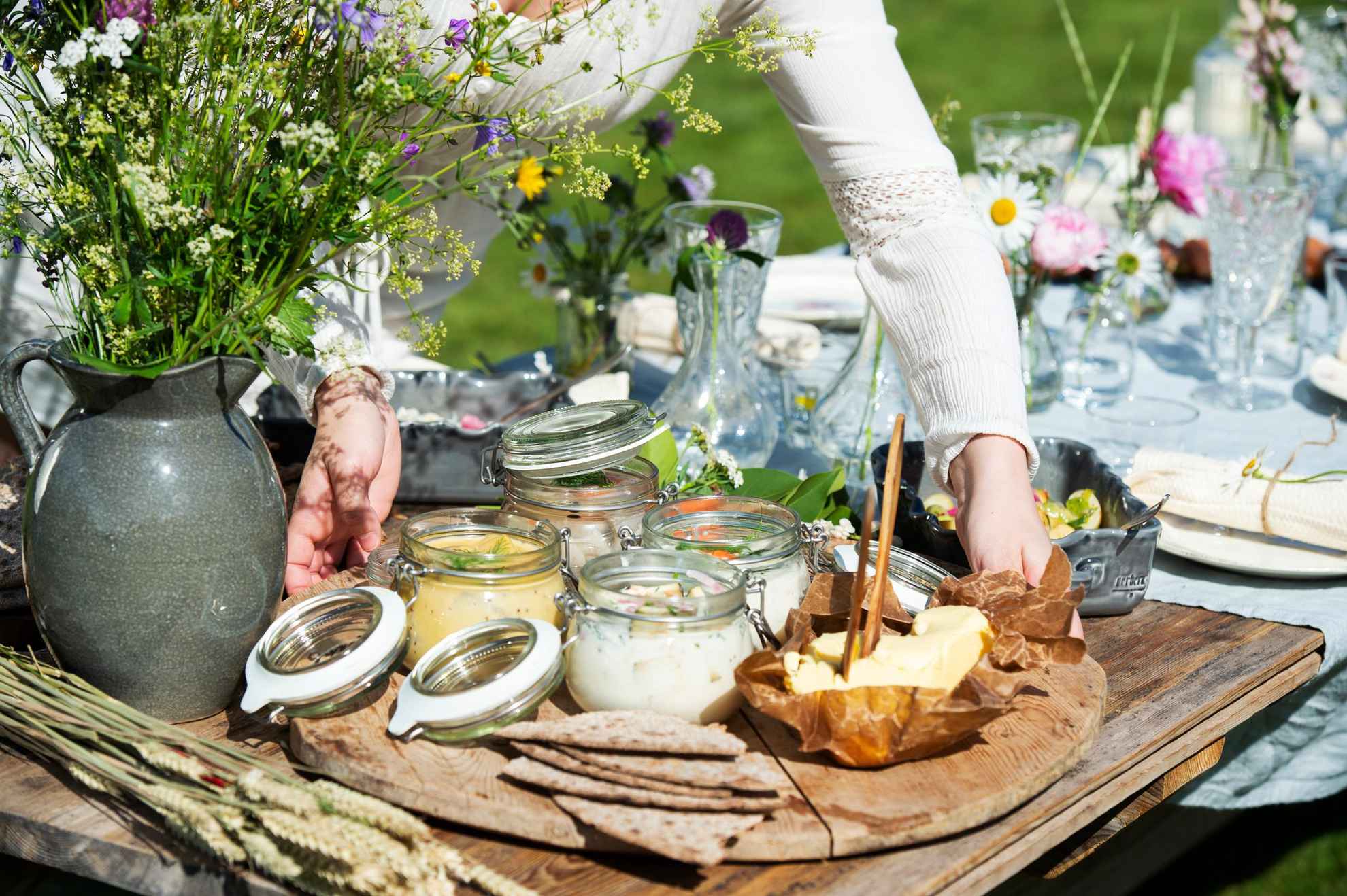 A woman puts a tray with pickled herring, butter and bread on a table set for midsummer lunch. Next to the tray is a vase with wild flowers.