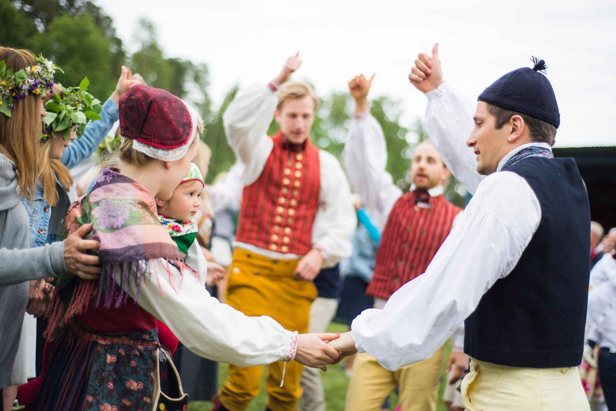 Men, women and children are holding hands and dancing, wearing traditional folk clothing.