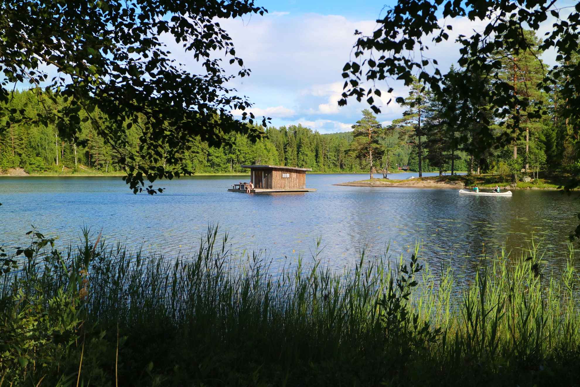 A cottage floating in a lake at Naturbyn and someone passing by it in a canoe. The lake is surrounded by forest.