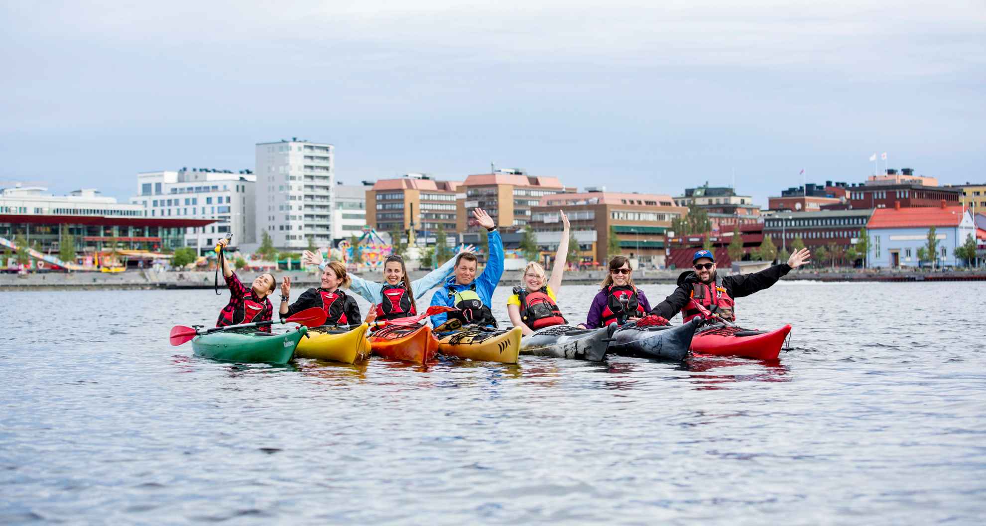 A group of seven people are kayaking. They are gathered and facing the camera in the sea in front of Luleå city.