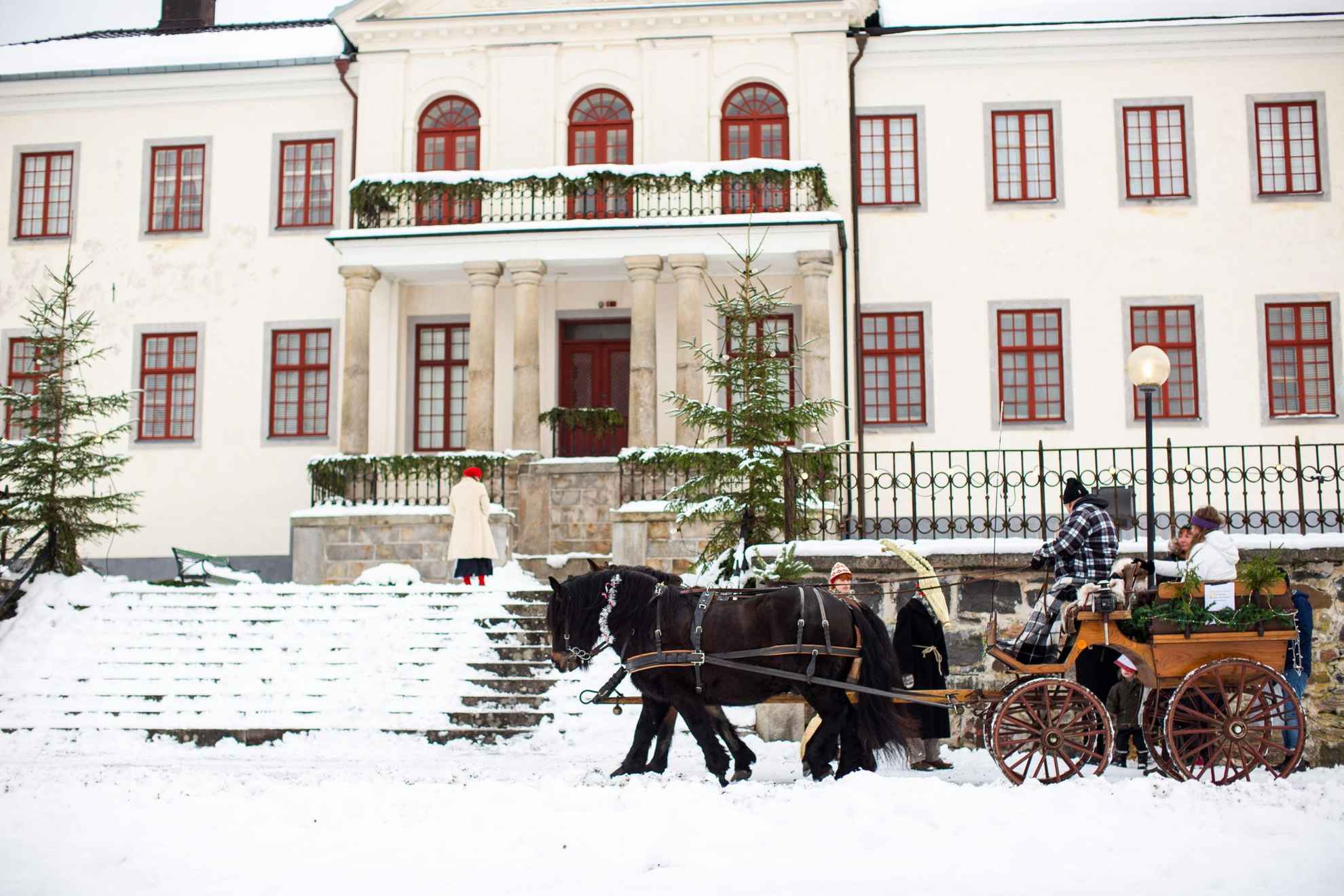 A horse and carriage stand in front of a large white house during winter. There is snow on the ground, a few people standing next to the carriage and a few sitting on it.
