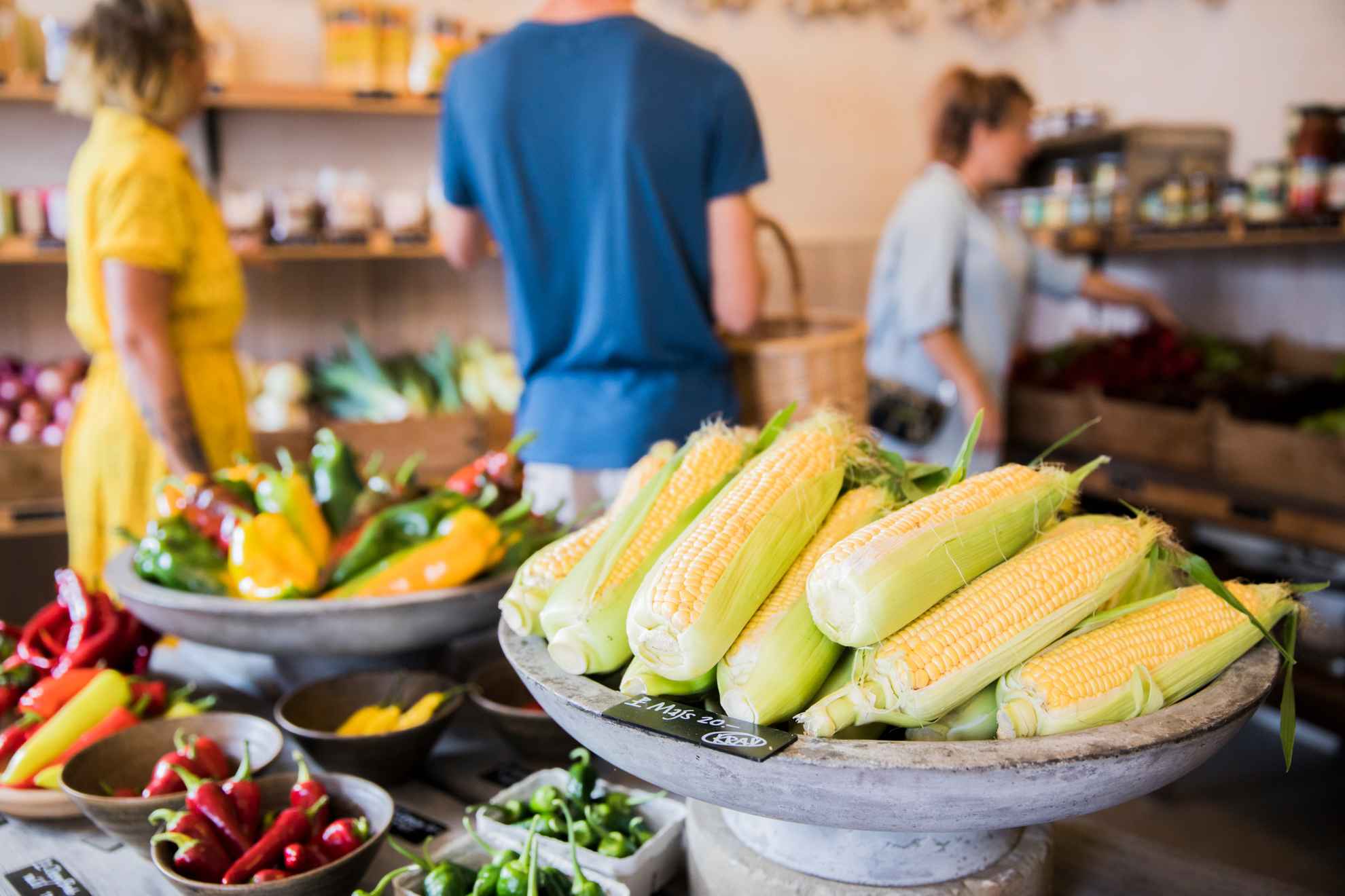 Close-up on corn-cob and a variety of pepper in a farm shop. Three persons is seen blurry in the background.