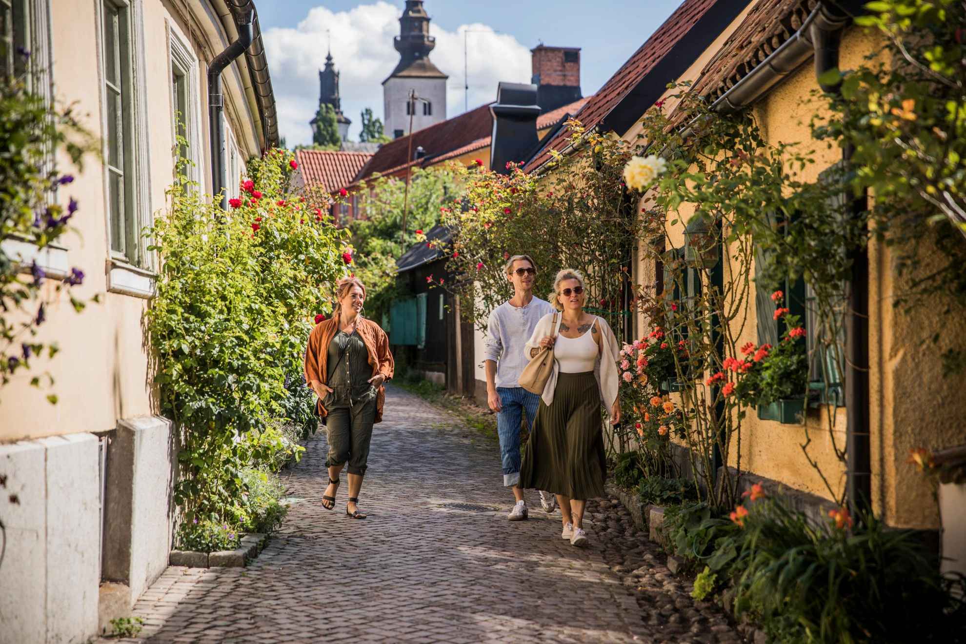 A man and two women strolling down a small cobblestone street in Visby. Flowers are growing in front of the old houses.
