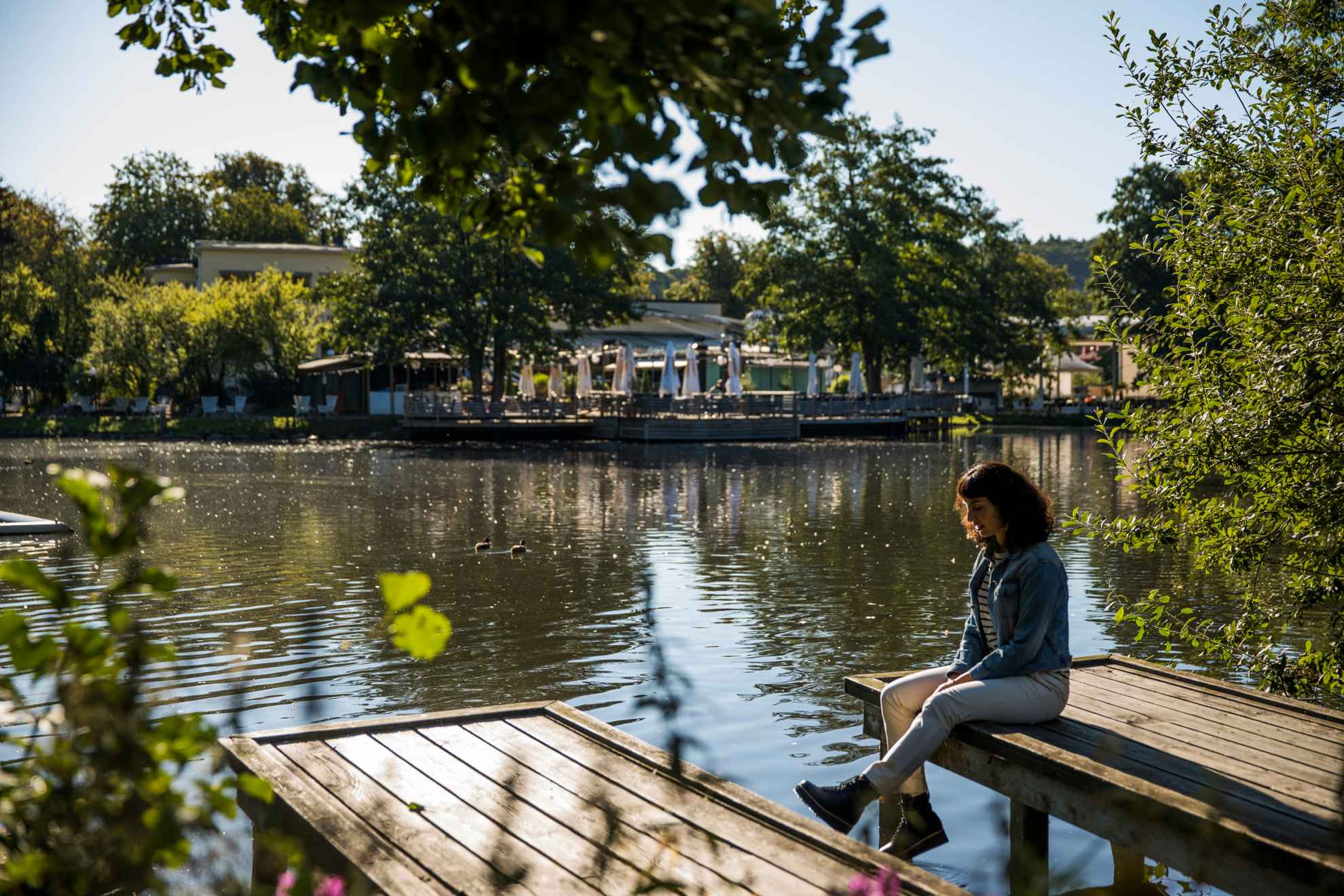 A woman sitting on a jetty next to a pond surrounded by greenery in a park. In the background you see an outdoor restaurant.