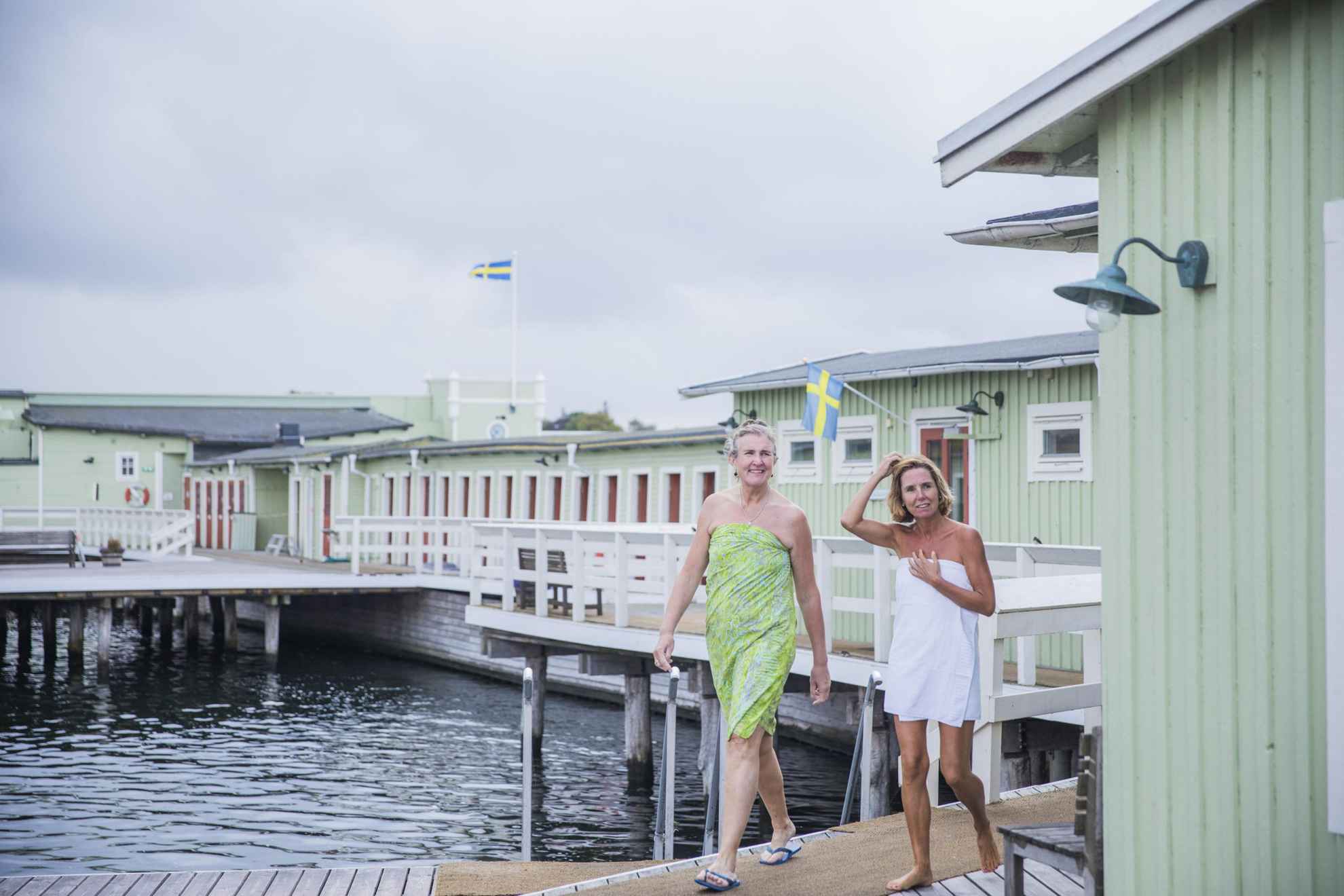 Two older ladies is going for an outdoor swim. They are both wearing towels and are walking on the decks of the open-air cold bath house of Ribersborg. The sky is grey and there is no sunlight visible.