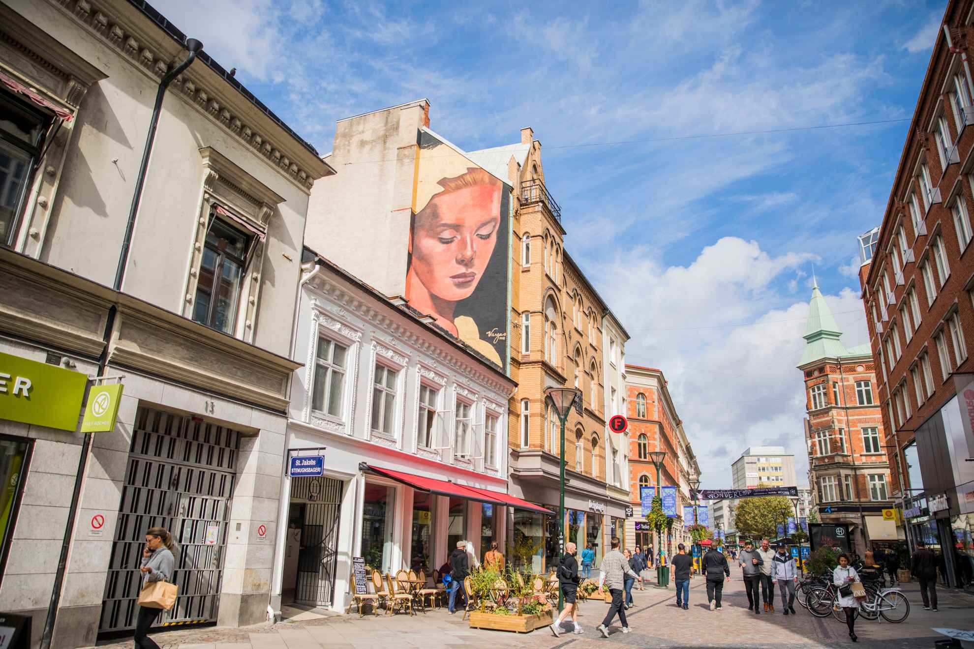 A pedestrian street with stone houses on each side. People strolling down the street. A mural painting on a house showing a the face of a woman.