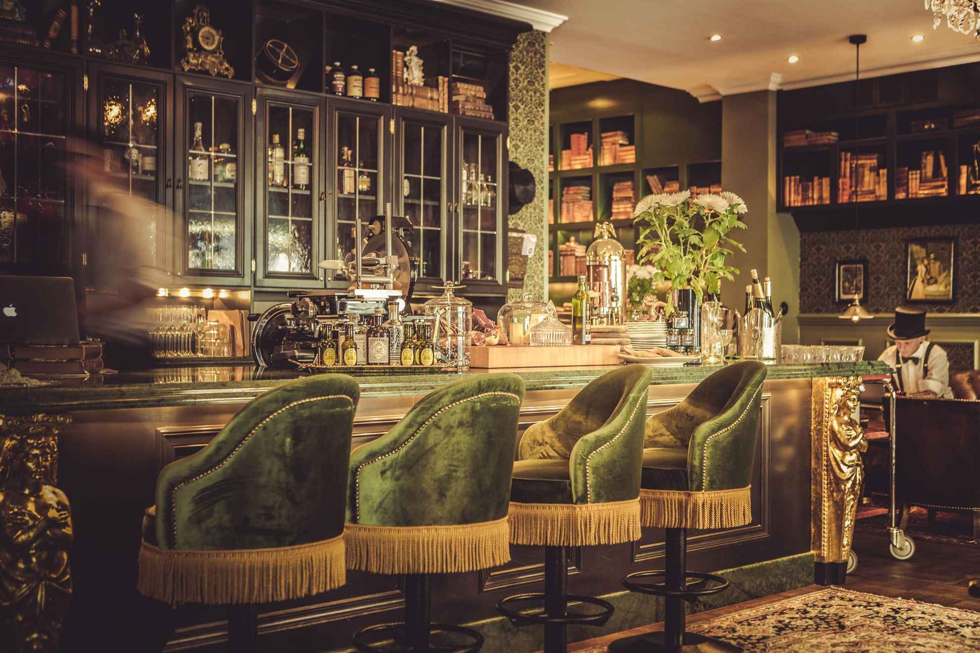 The bar at hotel Pigalle, with green velvet chairs, wooden shelves and golden details. Someone moving behind the bar.
