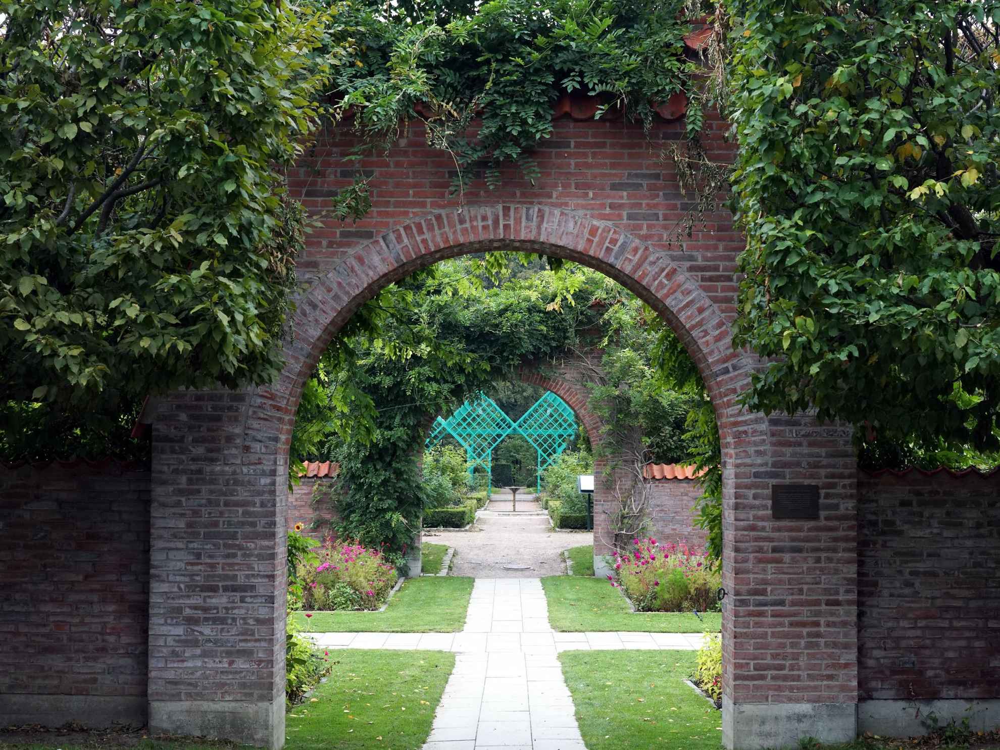 Inside a park, a walkway through two arches covered with greenery. There are beautiful plants and flowers. At the end of the road you see a blue square construction.