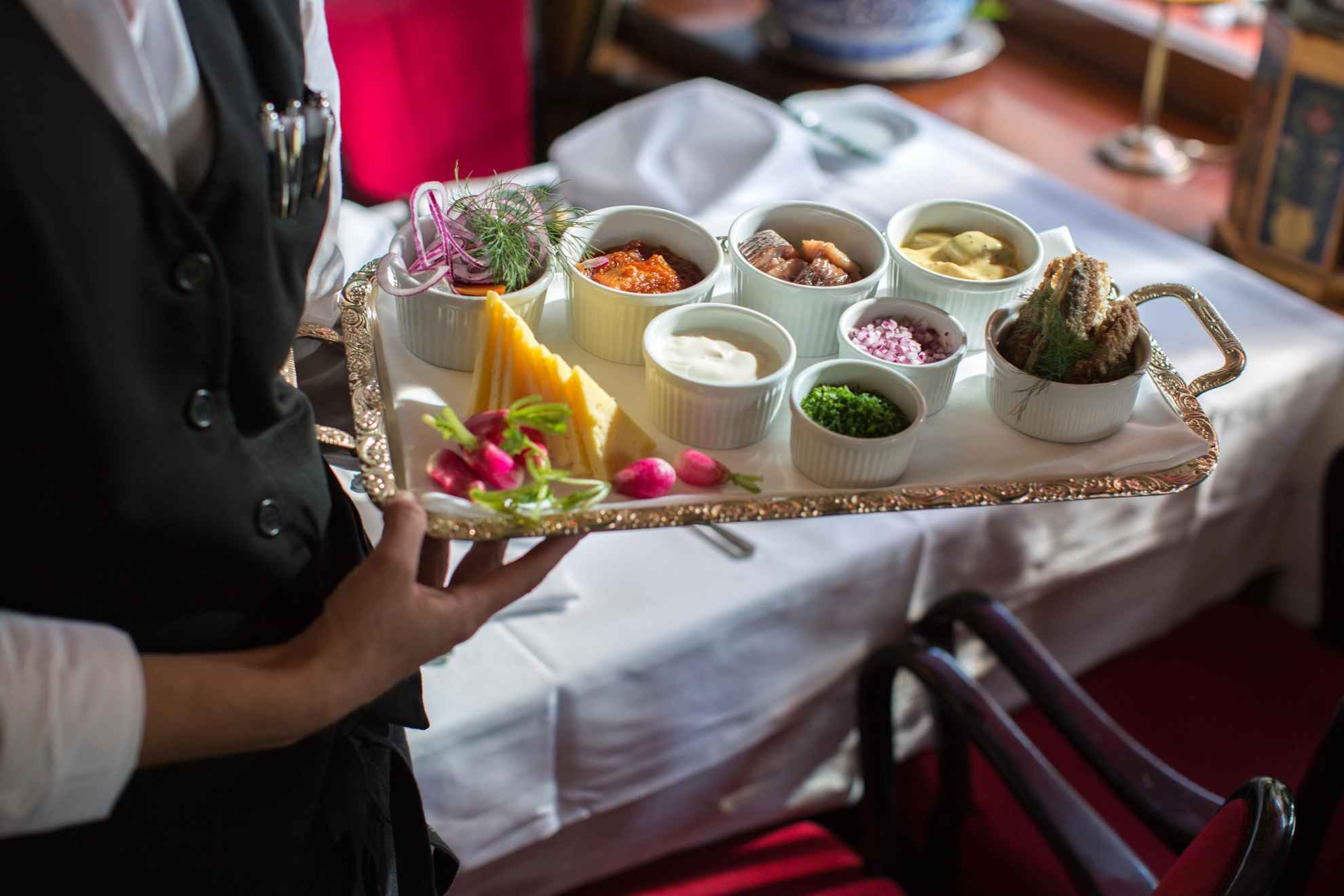 A waiter holds a tray with small bowls of chive, red onion, sour cream and herring.