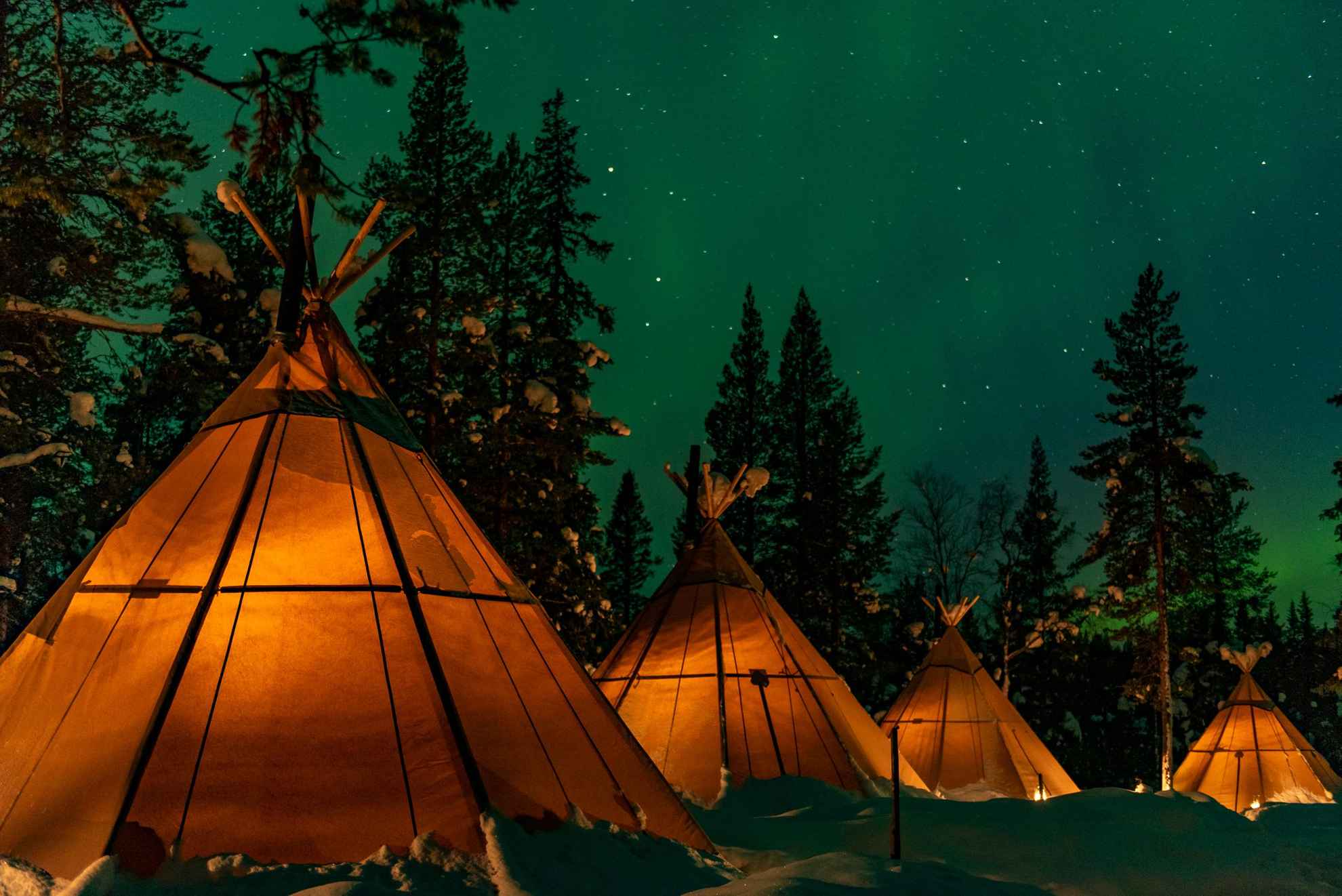 Four lavvu tent at a nature camp in Lapland. It’s a starry night in a snowy landscape and the light glows in the tents.