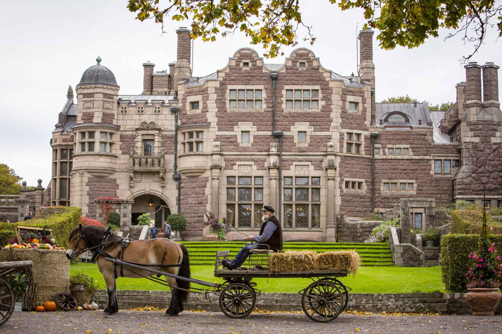 An elderly man is driving a horse and carriage with a couple of hay bales. They stand in front of a castle and beside you see a market stall with vegetables.