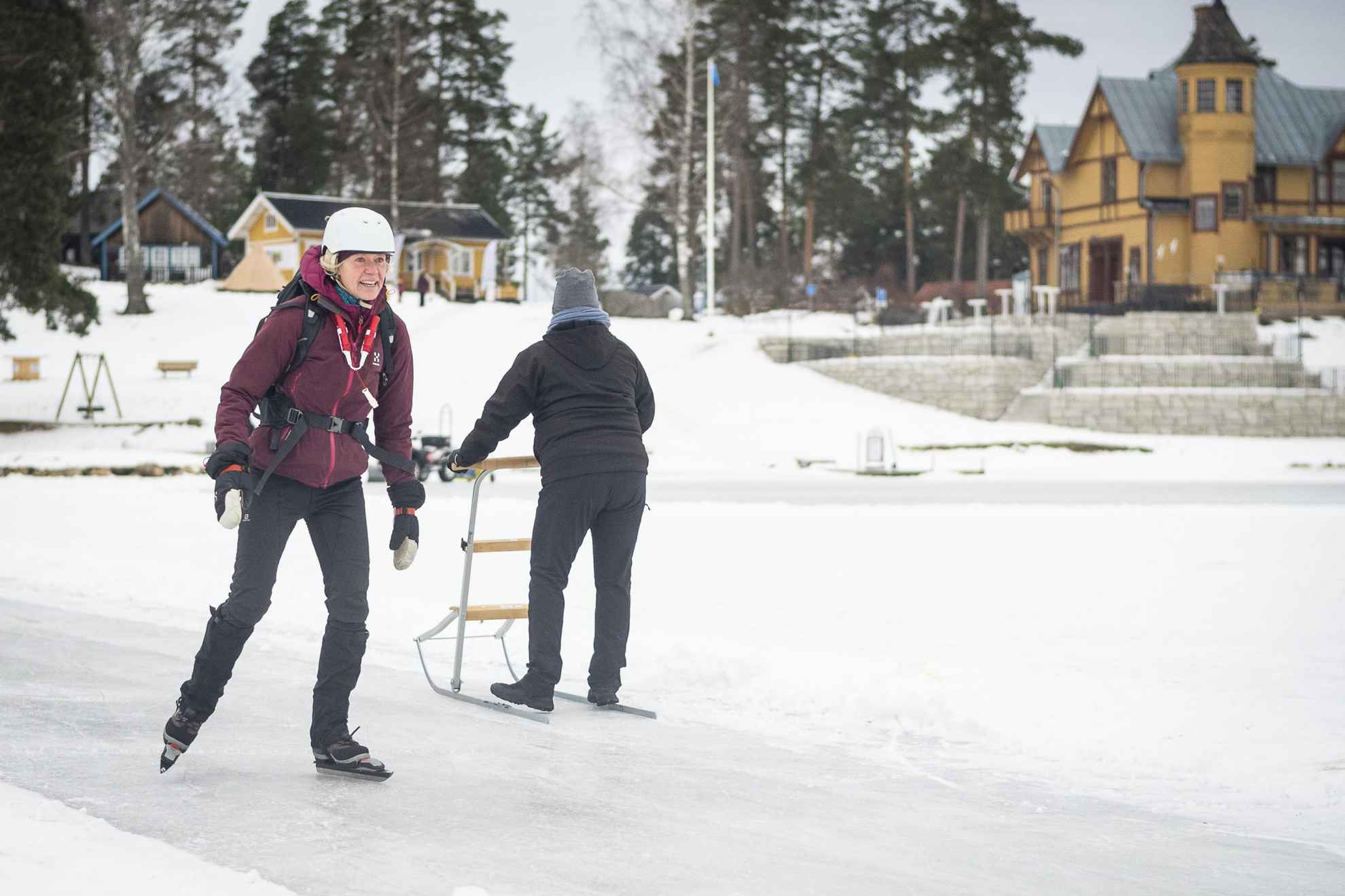 A woman is long distance skating while another person behind her is pushing a sled on the ice.