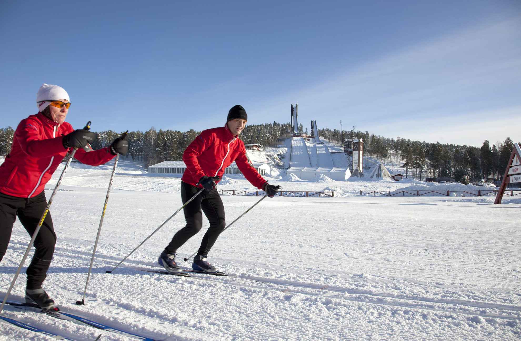 Two people in red jackets are cross-country skiing in prepared tracks.