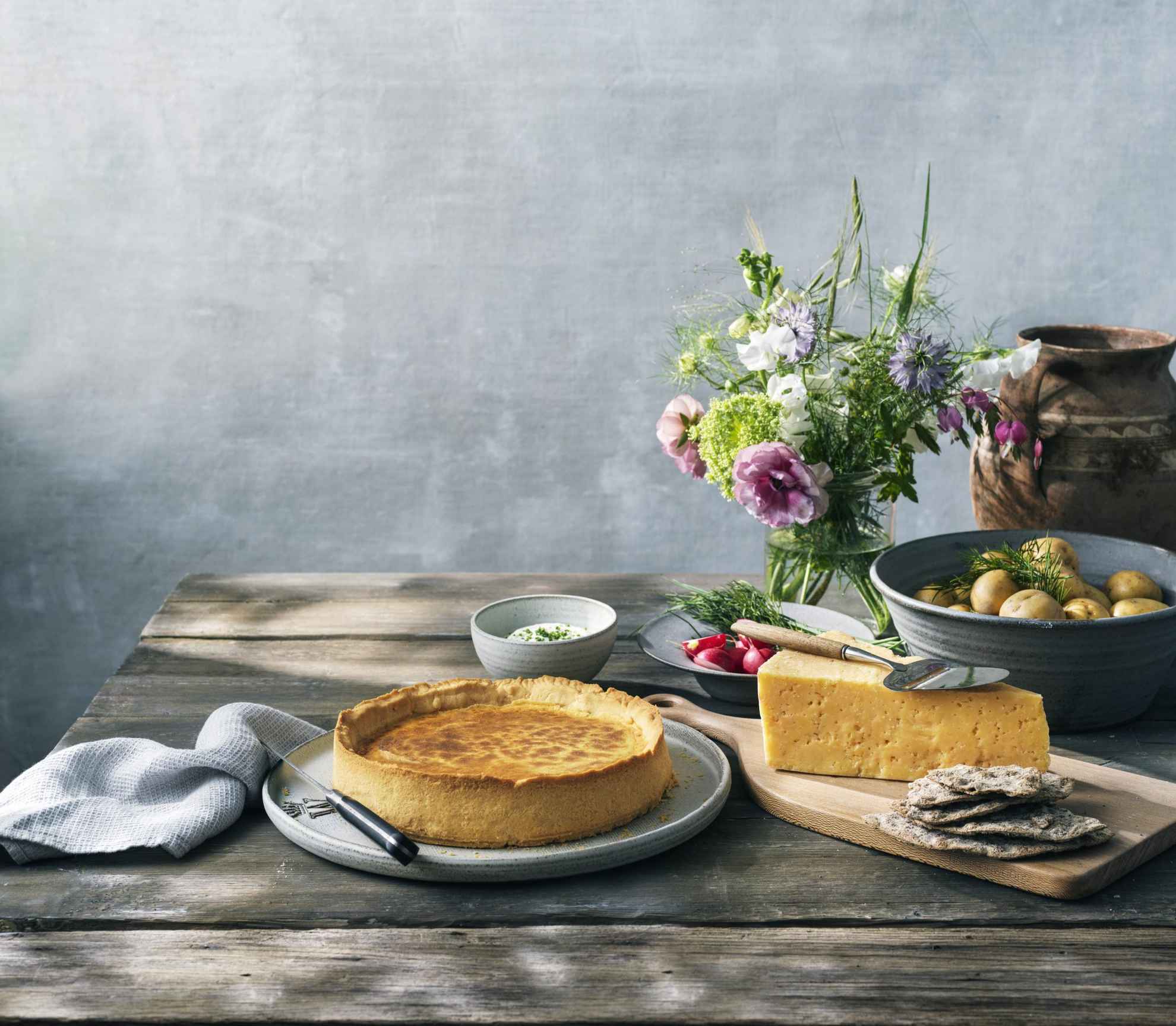 A wooden table with Västerbottenpaj quiche, Västerbotten cheese, a bowl of potatoes and a vase of flowers.