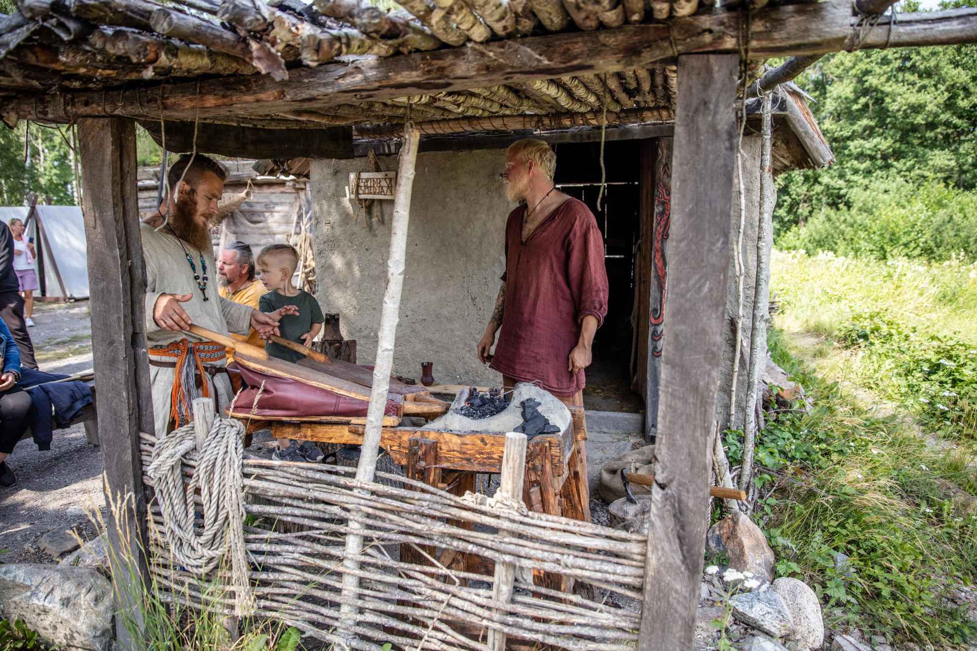Two kids watch as two men, dressed in historically typical viking clothing, work by a hearth.