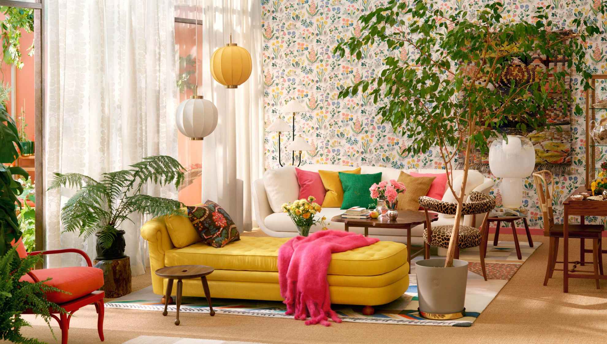 A living room with a beige sofa with colourful cushions at the back and a yellow chaise longue in the centre of the room. To the right of the chaise longue is a large green floor plant. The sheer white curtains let in natural light.