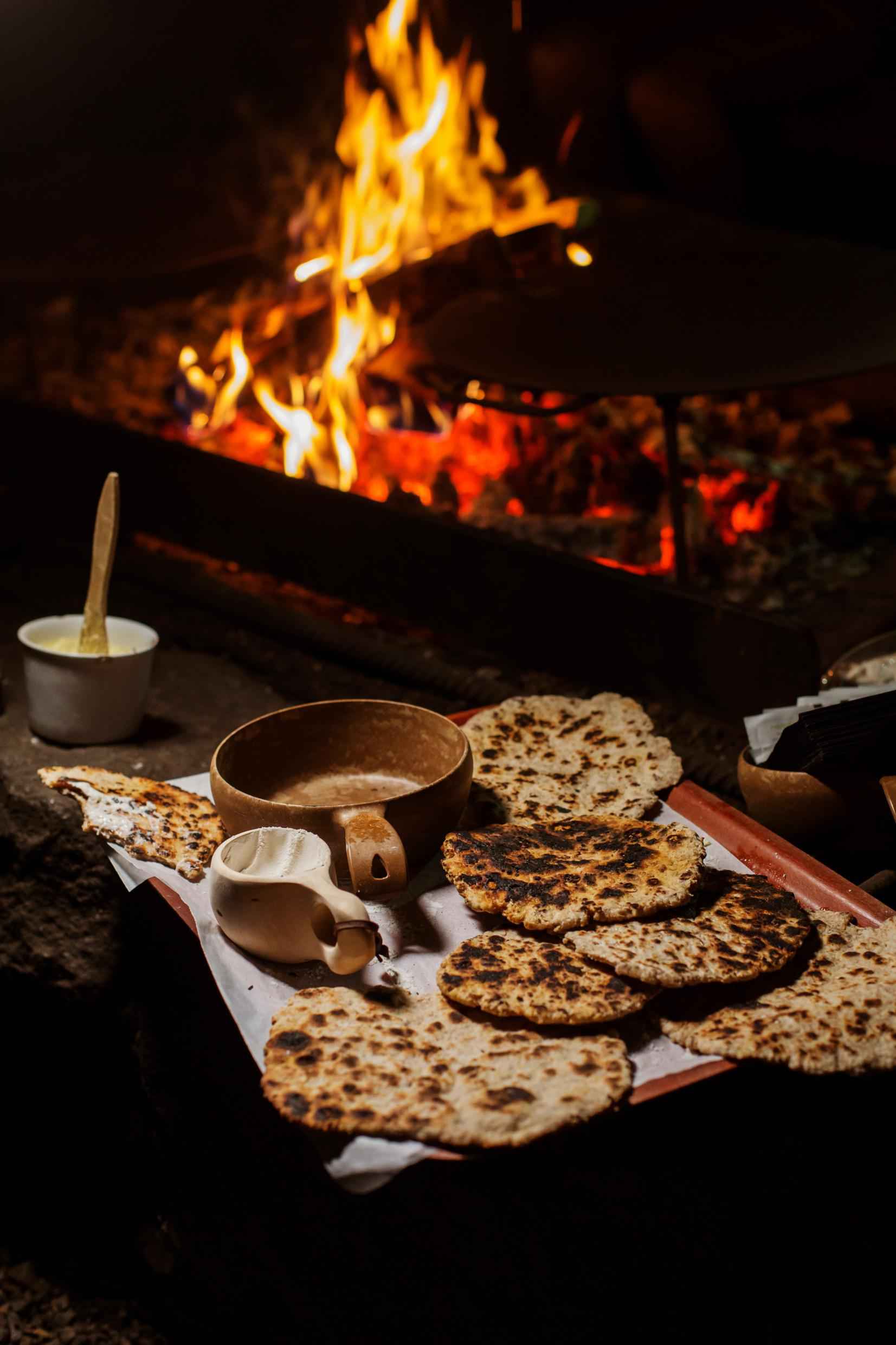 A tray with freshly baked flatbread and two wooden cups. A jar of butter next to the tray, and a campfire behind it.
