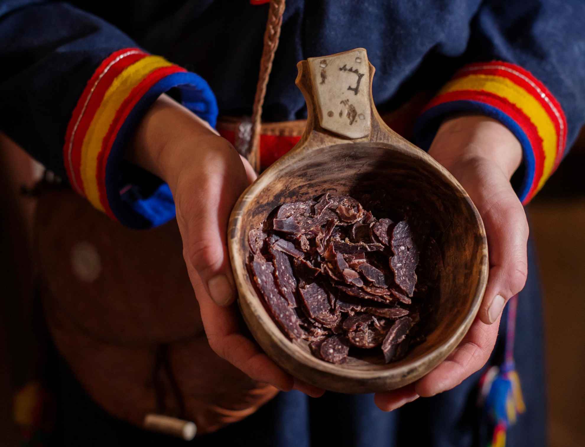 A person dressed in Sámi clothes holding a wooden cup filled with pieces of dried reindeer meat.