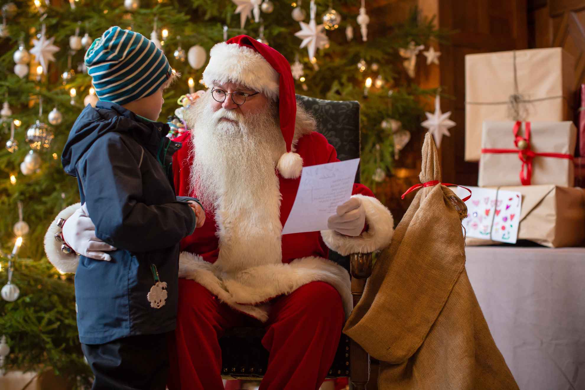 Father Christmas, an old man with white beard wearing a red gown sits in front of a Christmas tree talking to a child.