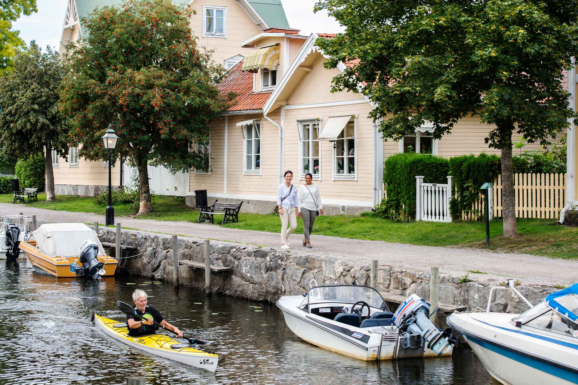 A man kayaking in a channal in a small town, where small boats lie. Two women walking along the channel.
