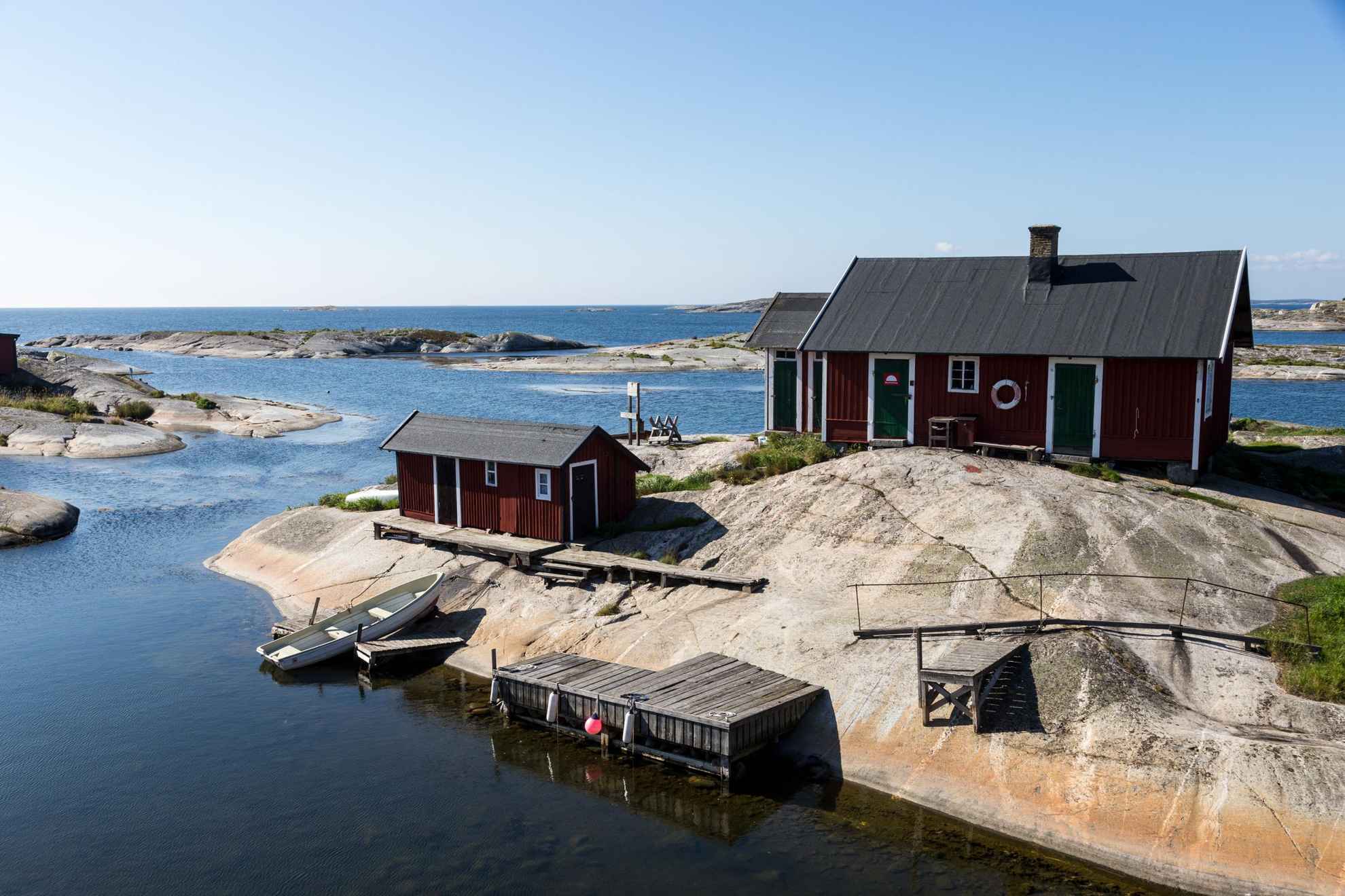 A house and a boathouse with a wooden pier sit on a cliff in the Stockholm archipelago. An empty boat sits on the ocean, halfway up on the cliff.