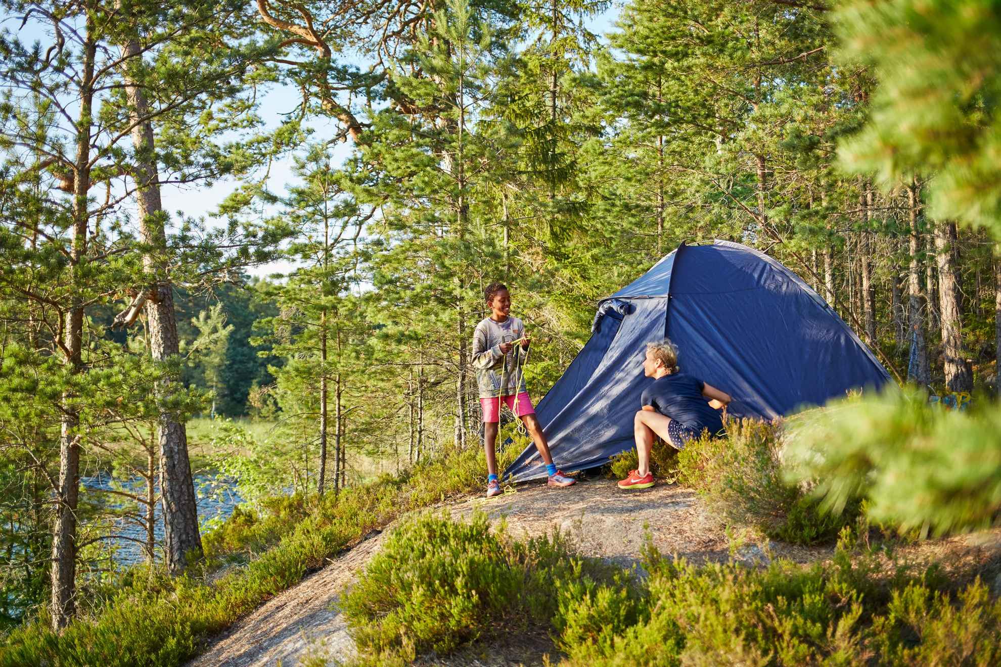 Two people in Sweden are by a tent in the woods next to a lake.