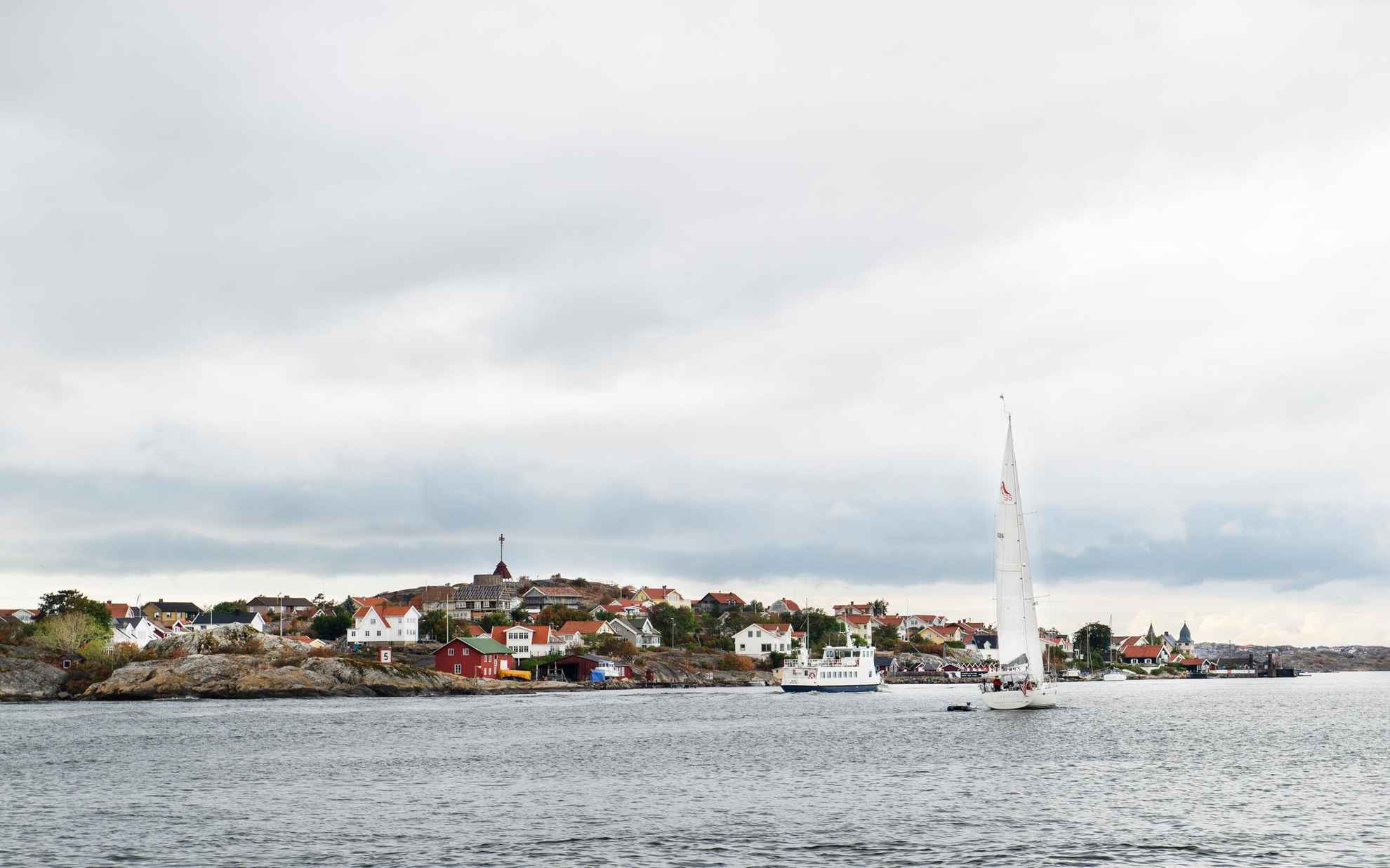 A small ferry and sailboat in the sea, passing by an island with many houses outside Gothenburg.