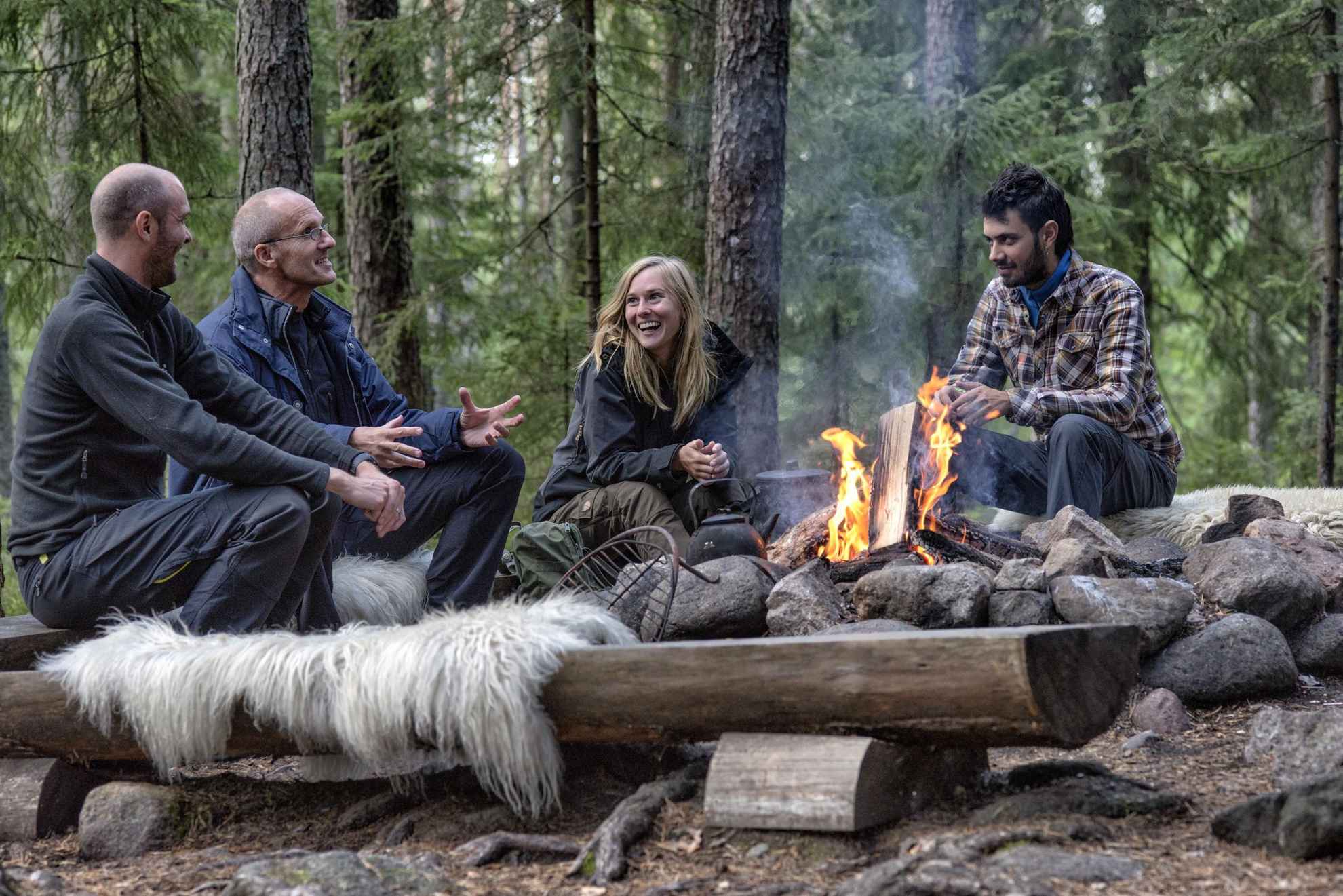 Four people sitting around a campfire in a forest laughing.