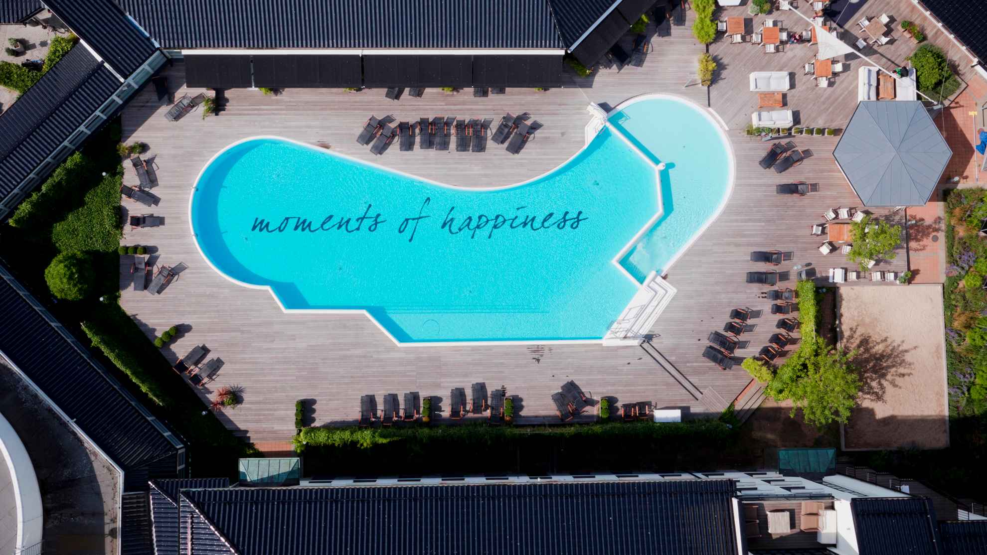 Outdoor pool with the inscription 'moments of happiness' at the bottom, surrounded by a wooden deck and sunbeds.