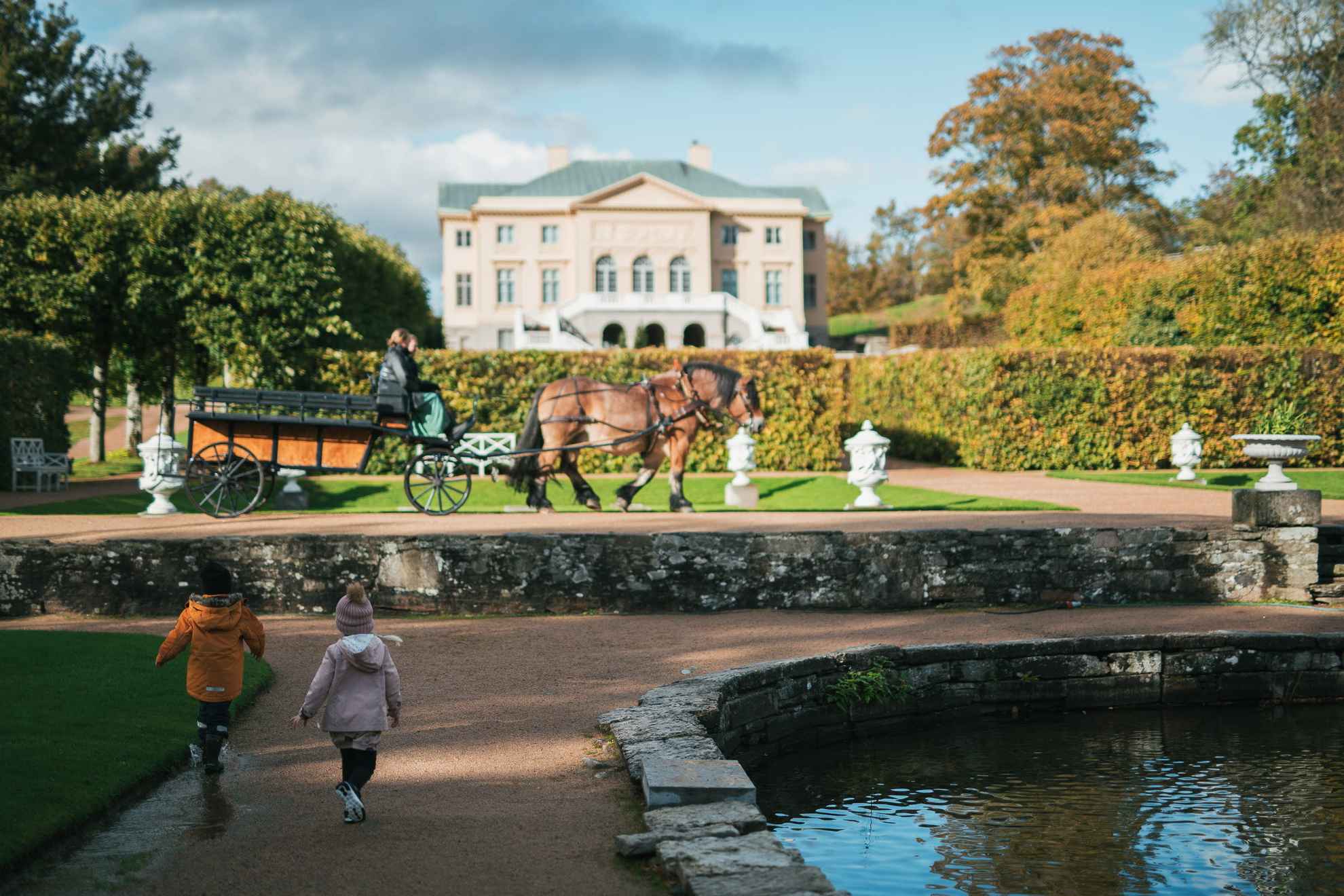 Children walk in a garden. There is a horse drawn carriage behind. A castle in the background.