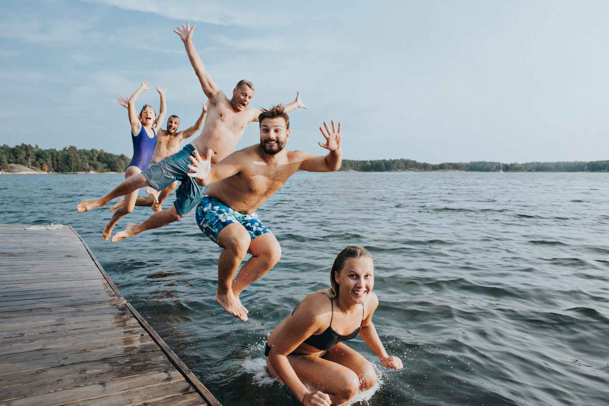 Five young people in swimwear are jumping into a lake from a wooden pier.