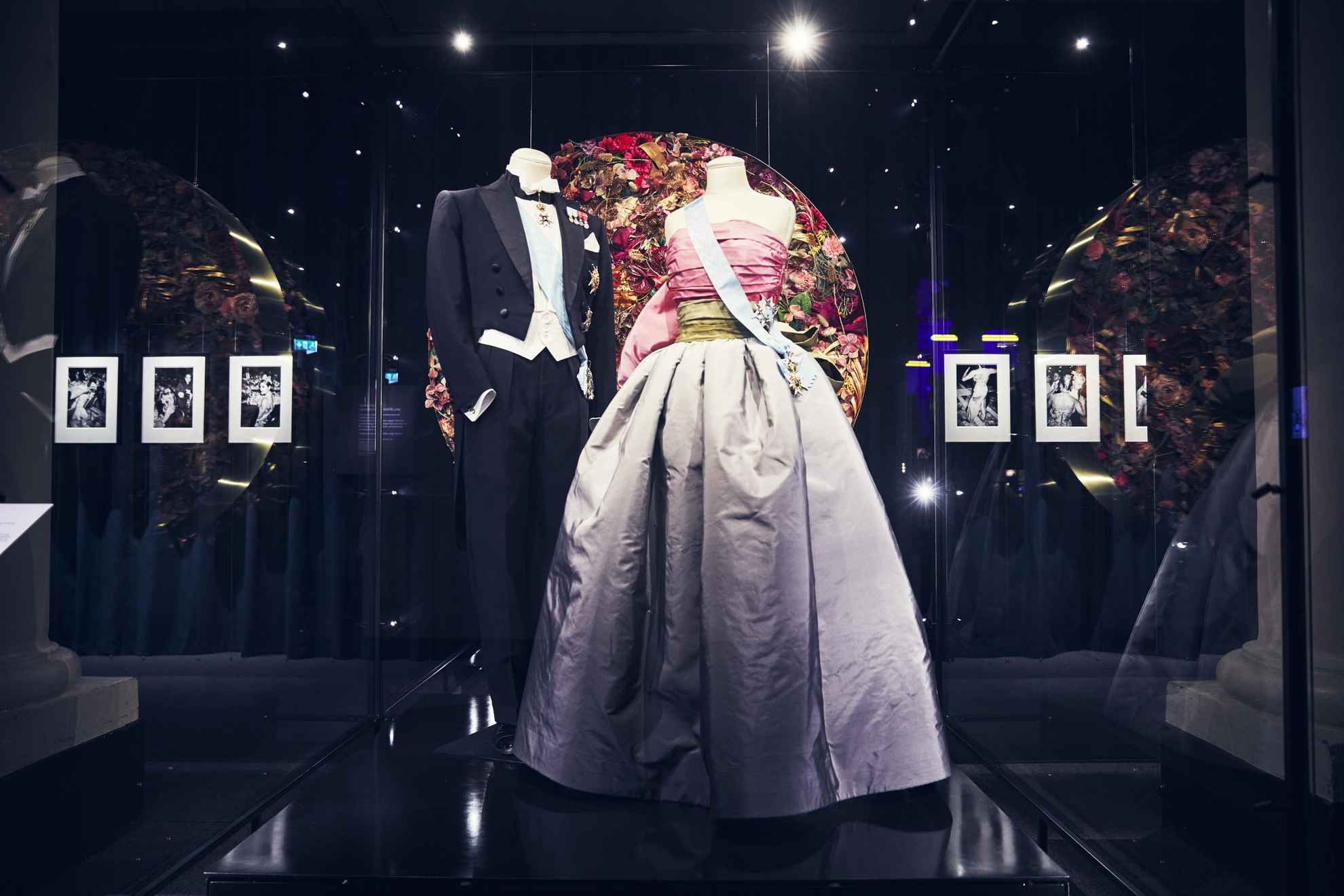 Two mannequins in an exhibition, one dressed in a tailcoat and the other in a purple and pink dress. Both wearing a blue ribbon.