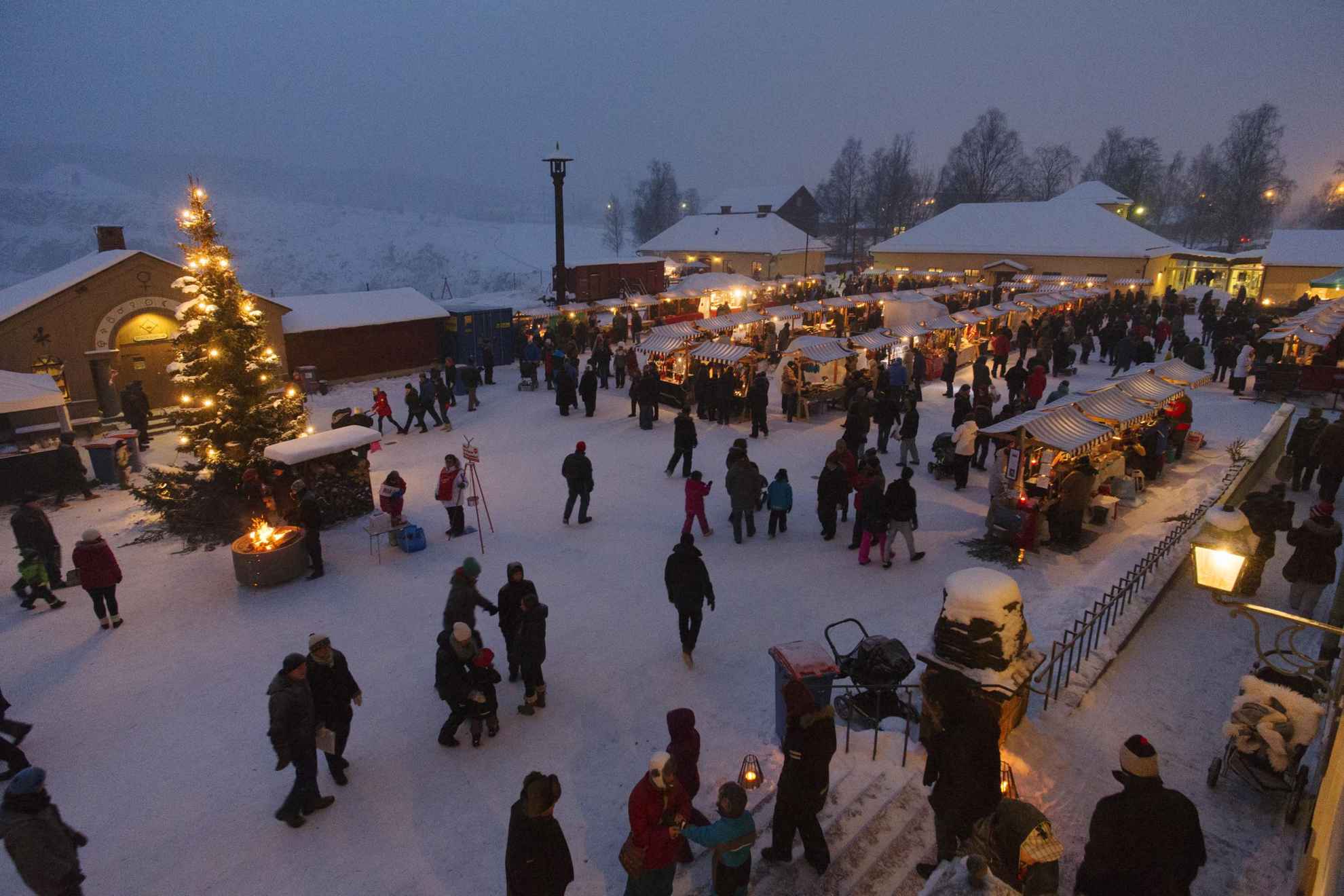 A Christmas market in the snow, with many stalls, people and a large Christmas tree by a little bonfire.
