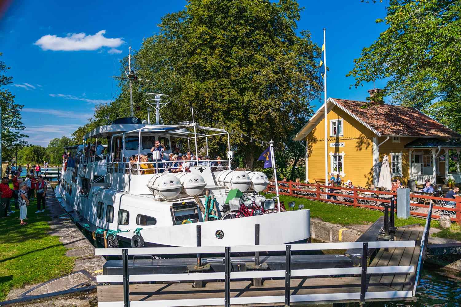 A large white boat passes by the yellow wooden house of Hajstorp hostel and café.