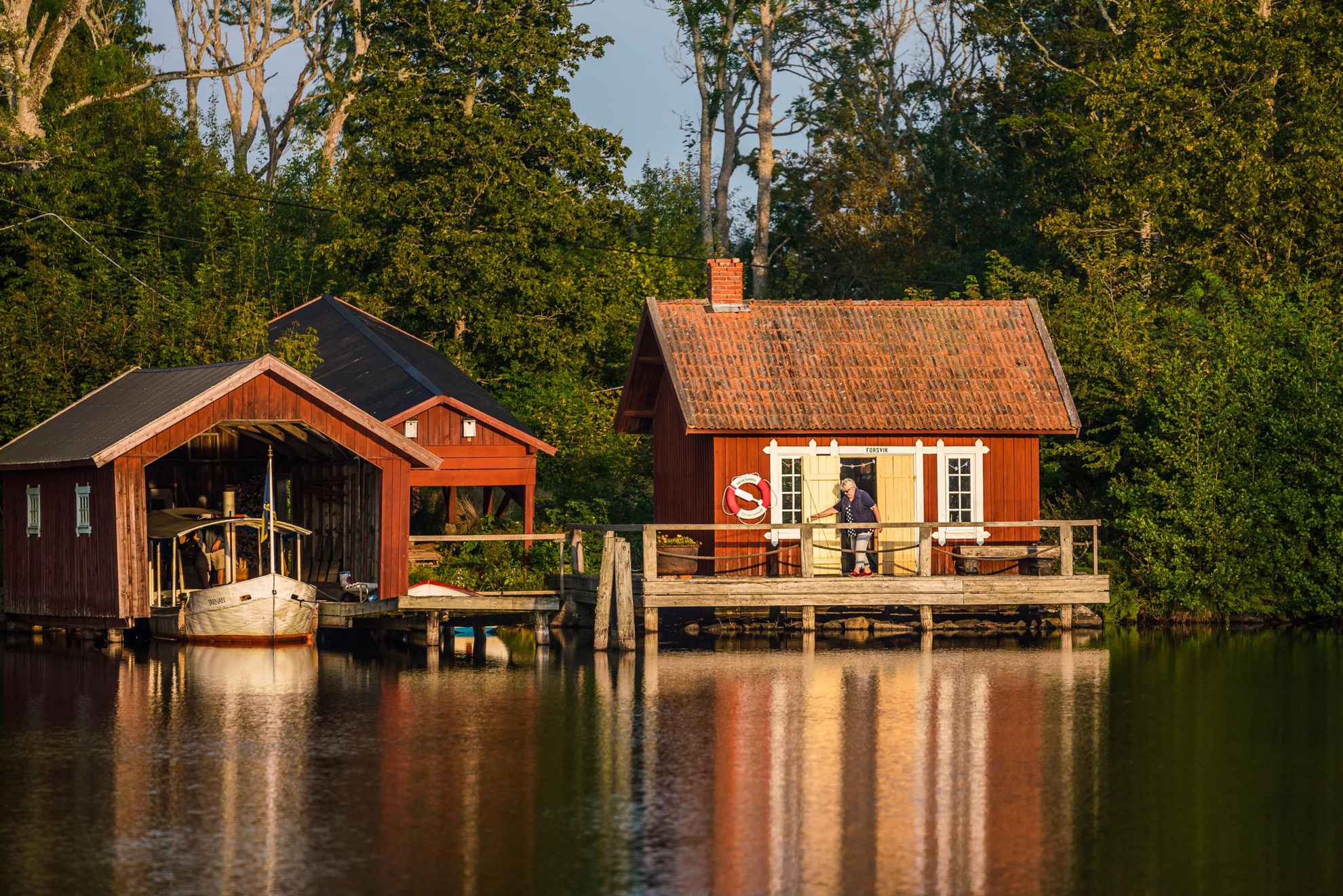 A woman is exiting a red wooden house that lies by the water. Next to the house is a boat house.