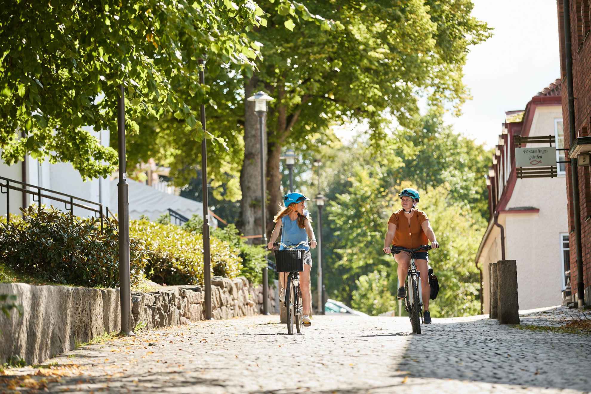 Two women are biking on a small cobblestone street. Houses and greenery along the street.