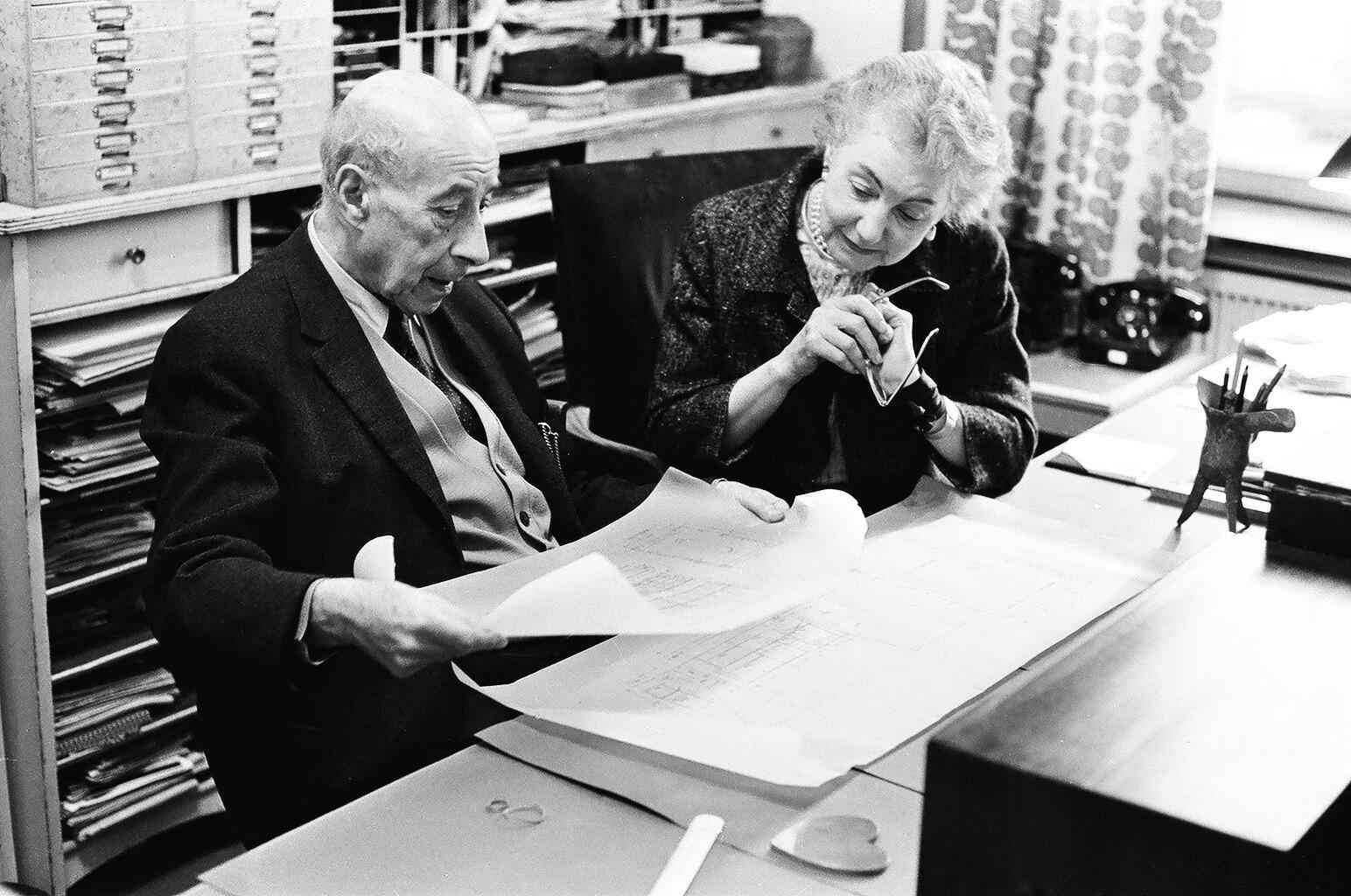 An older man and woman sitting at a desk looking at design drawings.