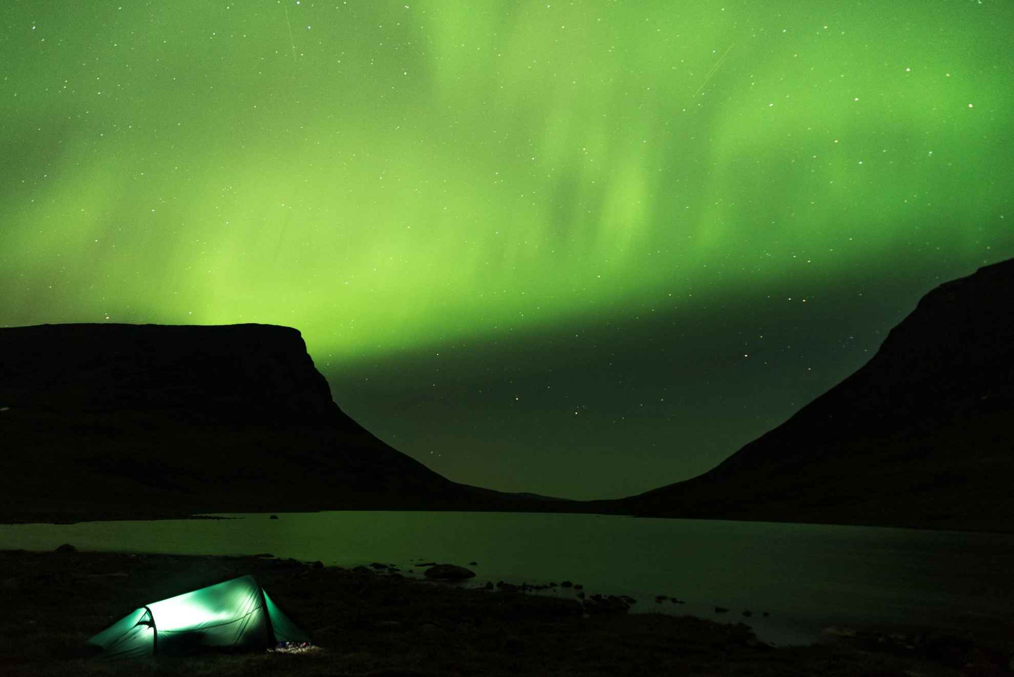 Northern lights are dancing above Lapporten mountains in Abisko. By Torne lake, a pitched tent is lit from inside.