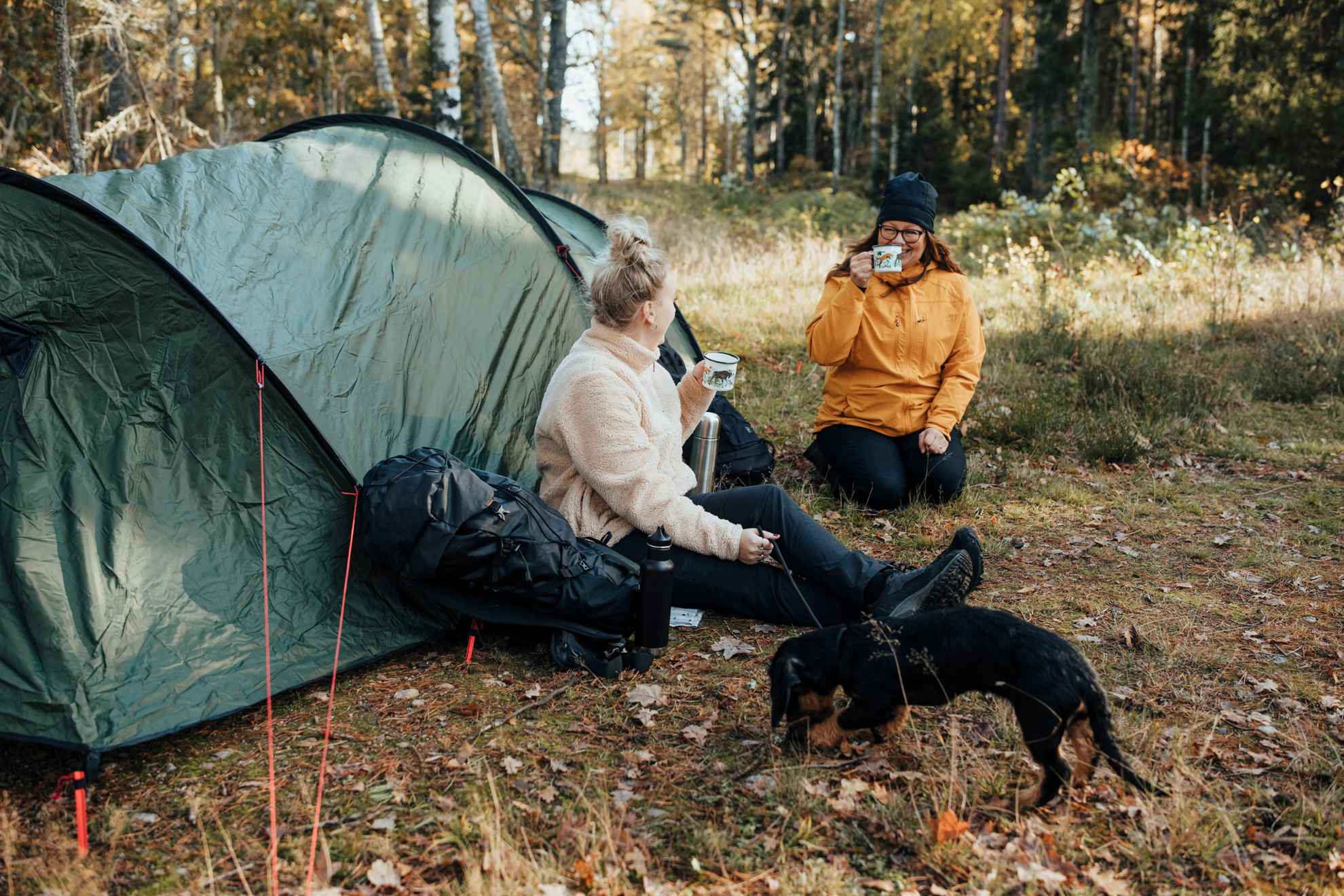 Two women in outdoor clothing sit on the leafy ground next to a tent and drink coffee from mugs. A black dachshund can be seen in the foreground.