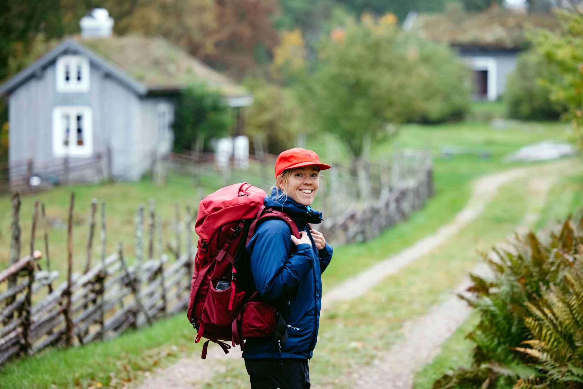 A woman in outdoor clothing and a backpack looks at the camera while standing on a hiking trail. A wooden croft is seen in the background.