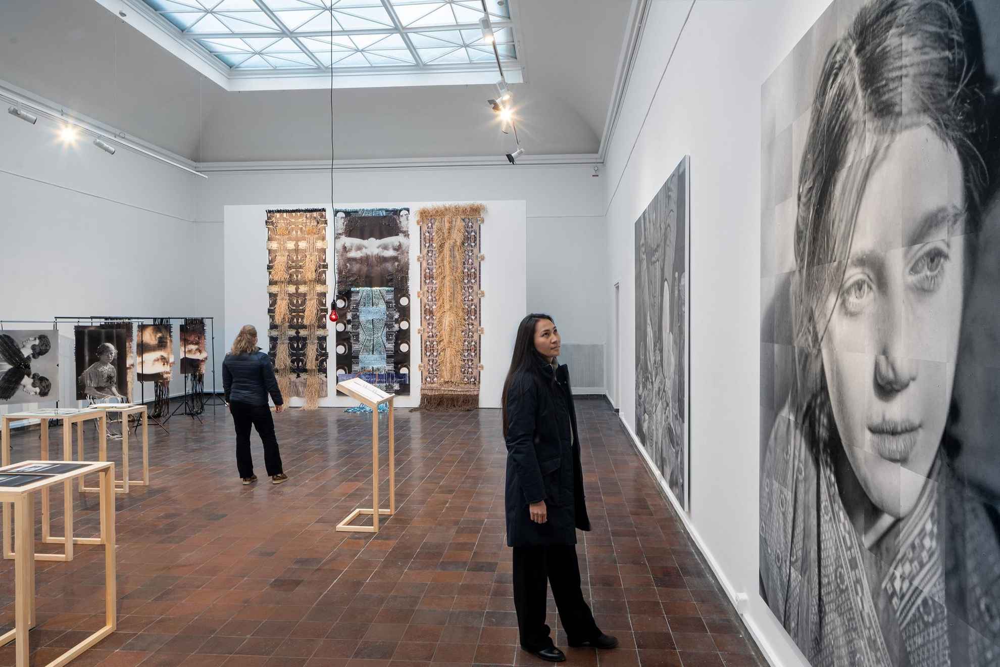 An installation at an exhibition. A woman looking at a large black and white image of a woman and in the background a woman is looking at a large artwork with photographs braided together with fragments into new structures.