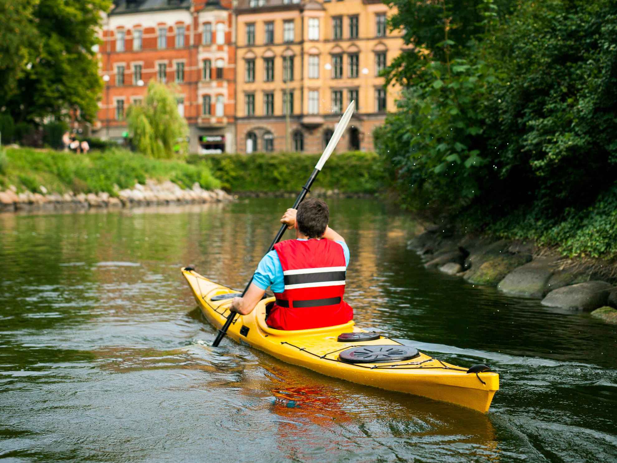 A man in a yellow kayak paddles in Malmö town. Houses in front of him and greenery along the sides of the canal.