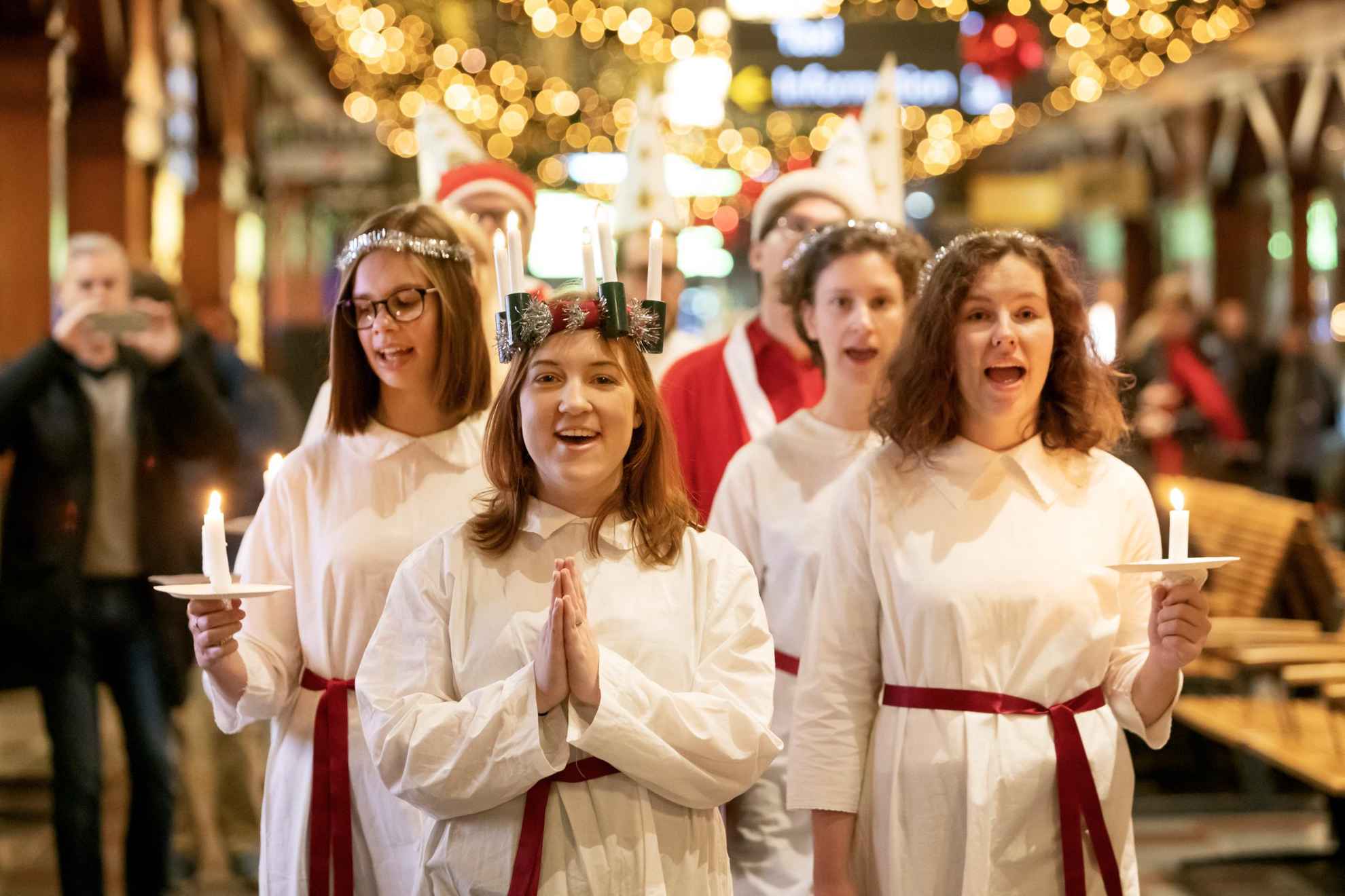 Girls dressed in white robes, holding candles and singing inside a train station.